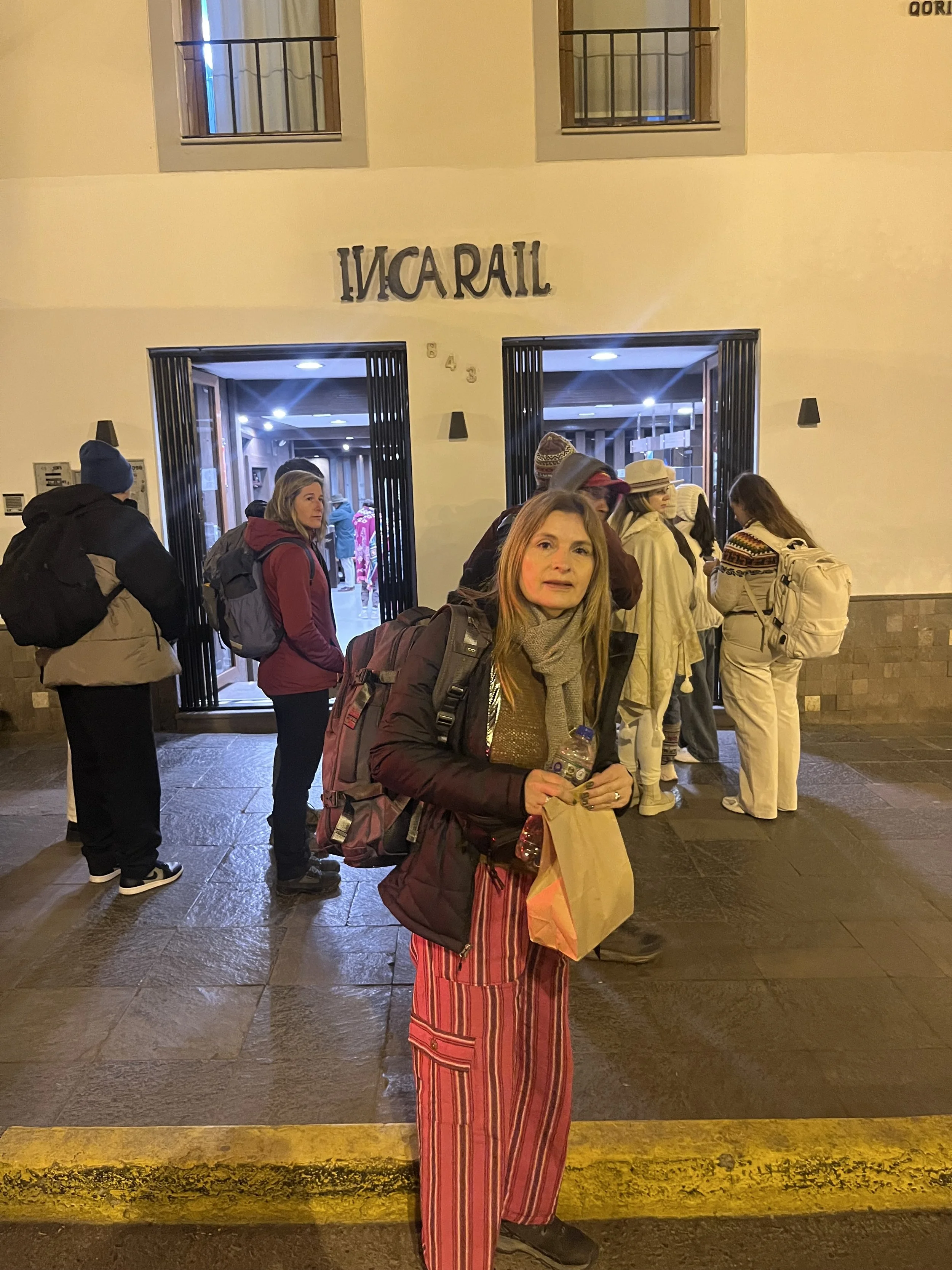Grupo de personas haciendo fila en la entrada de un edificio de noche, una mujer en primer plano con un saco oscuro, bufanda gris, pantalones a rayas y mochila, sosteniendo una botella de agua y una bolsa de papel.