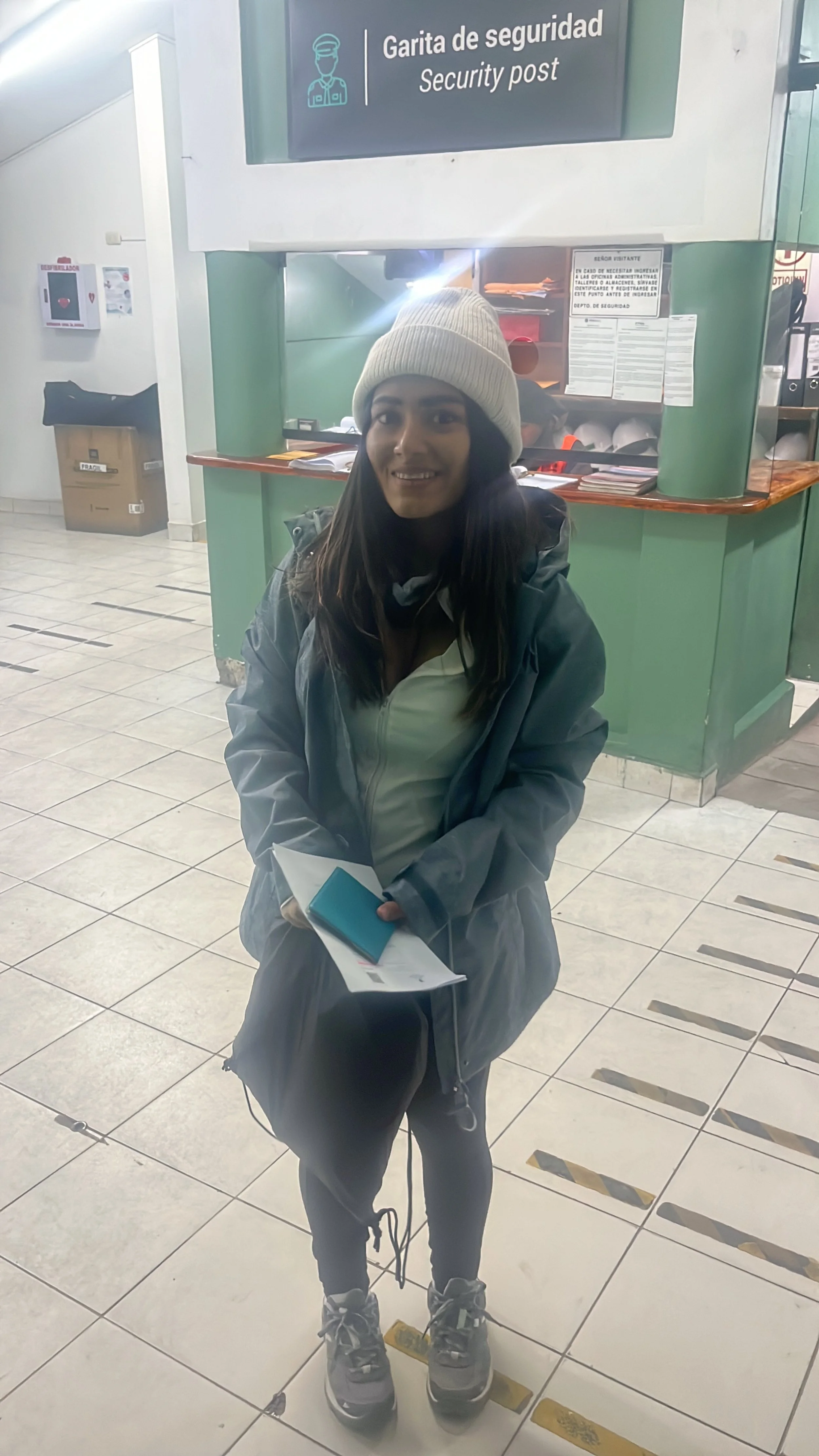 Persona joven con ropa de abrigo, gorro y botas, de pie frente a un puesto de seguridad en un edificio público, sosteniendo documentos y teléfono móvil.