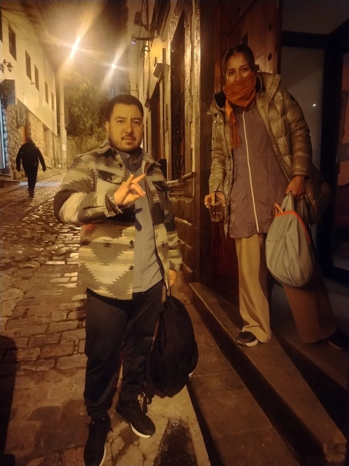 Dos personas en la calle callejón empedrado, una de ellas con camiseta de camuflaje y la otra con un abrigo y bufanda, con una mujer sosteniendo un vaso con bebida y mochila, en un ambiente nocturno.
