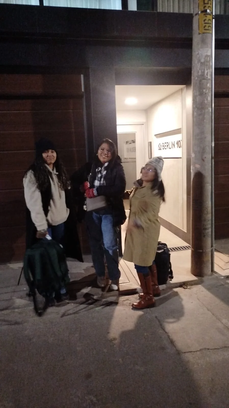 Tres personas con equipaje de viaje posando frente a la entrada de un edificio en la noche.