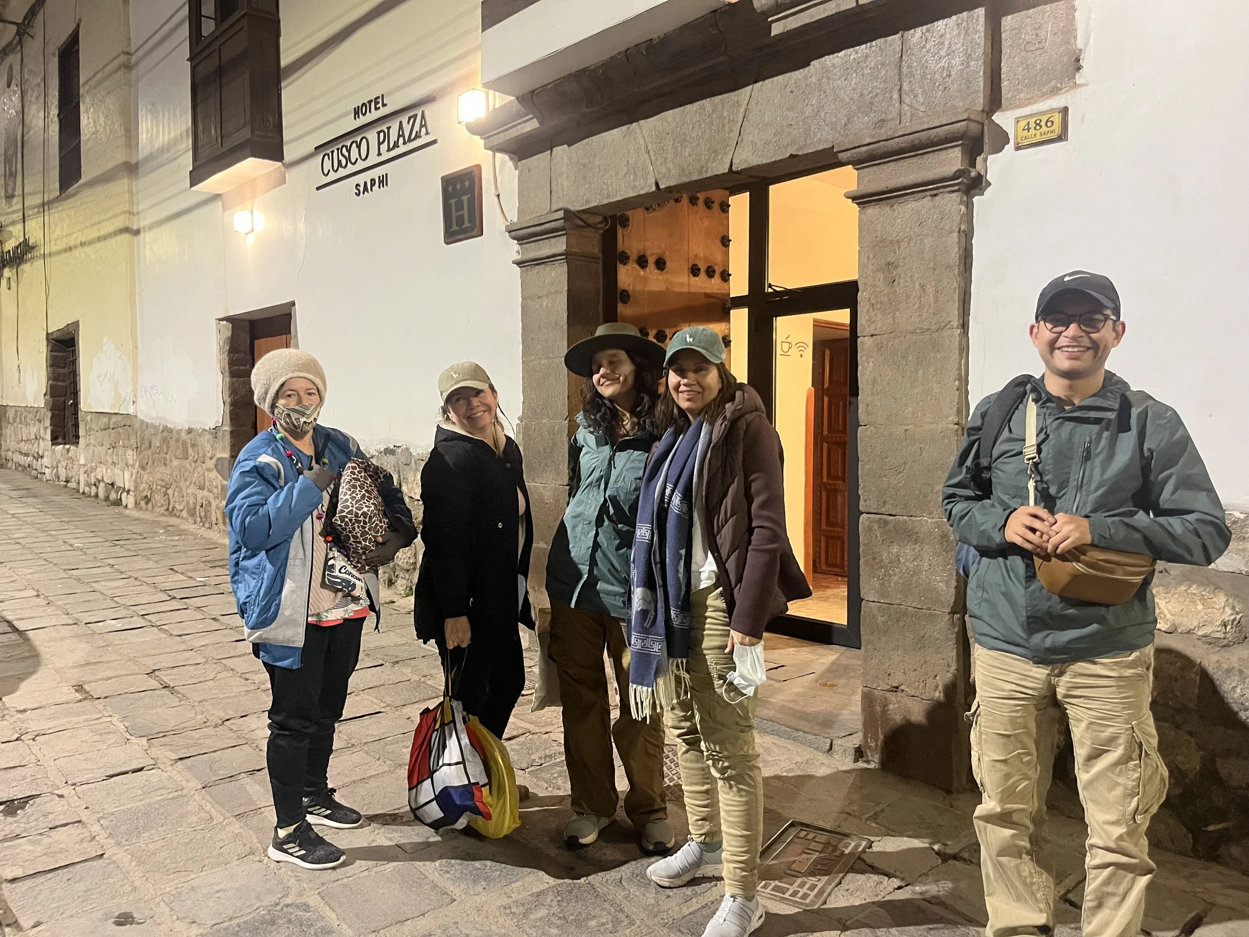 Grupo de cinco personas posando frente a la entrada de un hotel llamado "Cusco Plaza" en la noche, con calles de piedra y paredes blancas.