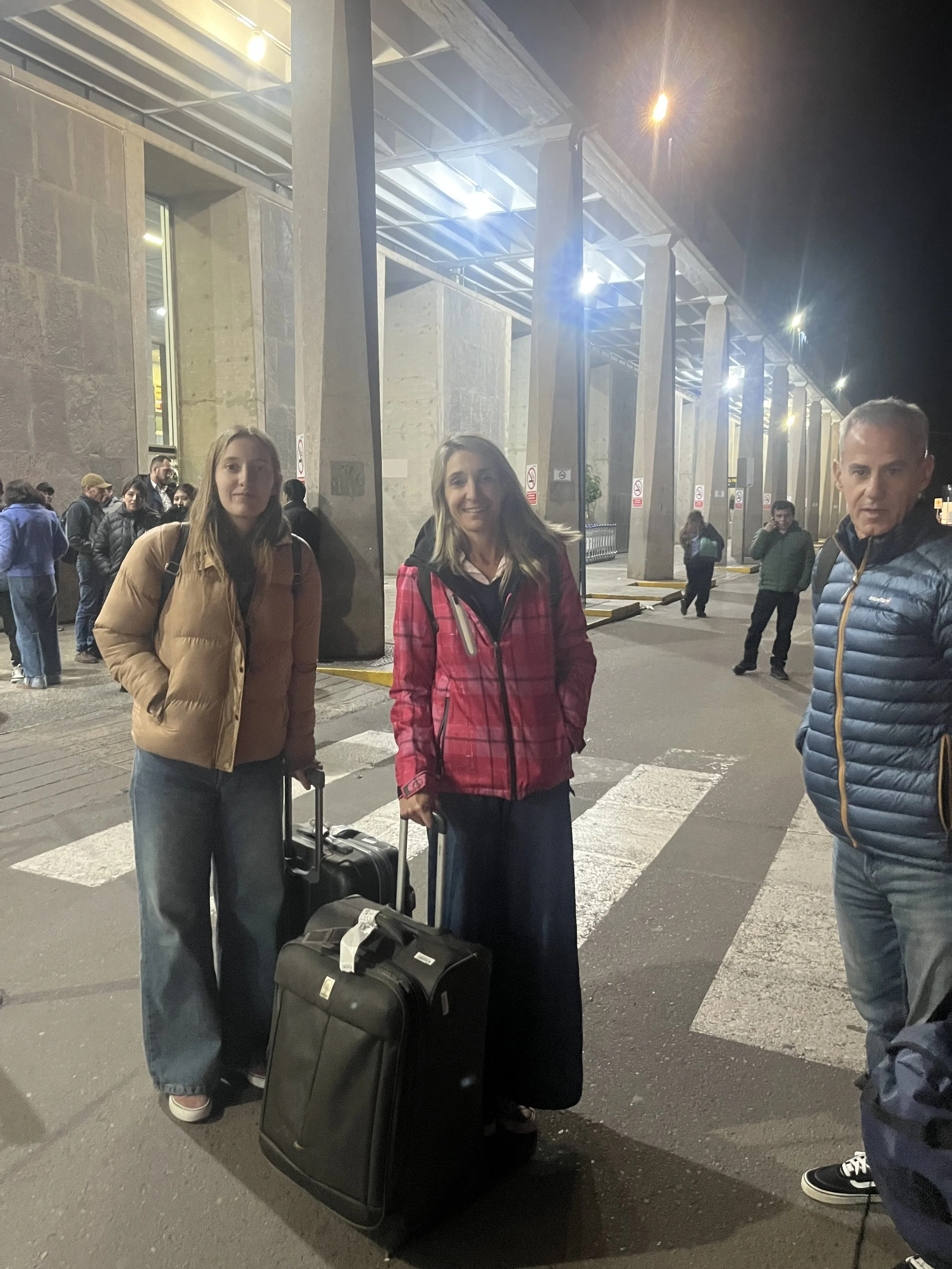 Tres personas con equipaje en una zona peatonal frente a un edificio con columnas, en la noche, con otras personas en el fondo.