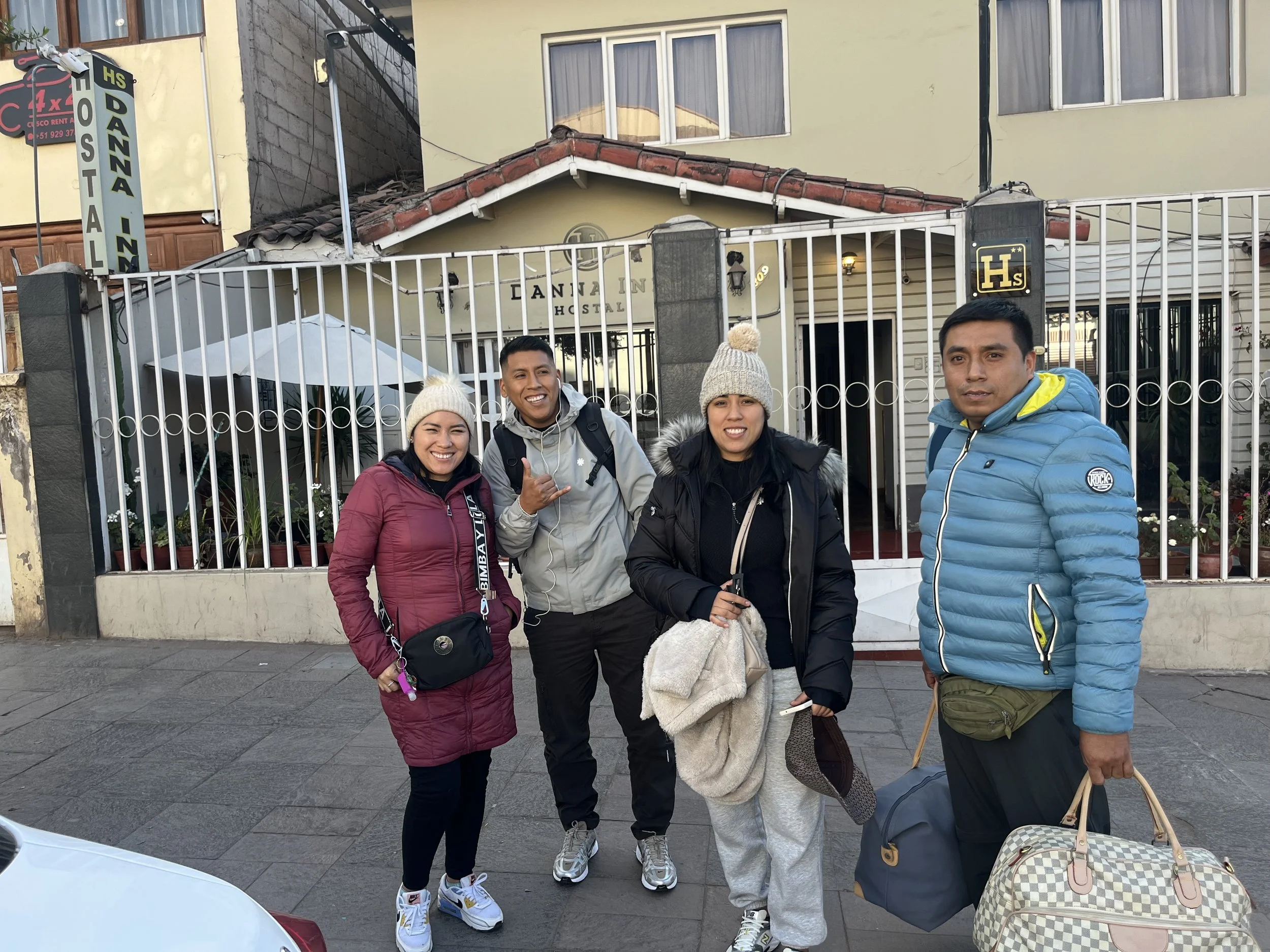 Cuatro personas sonriendo frente a la entrada de una hospedería, vestidos con ropa de abrigo y algunos con gorros, en la calle.