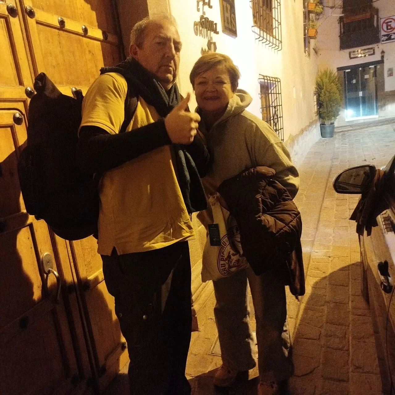 Hombre y mujer posando en la calle en la noche, ambos con ropa de abrigo, el hombre con mochila y mostrando un pulgar hacia arriba, en un entorno urbano con paredes con rejas y plantas en macetas.