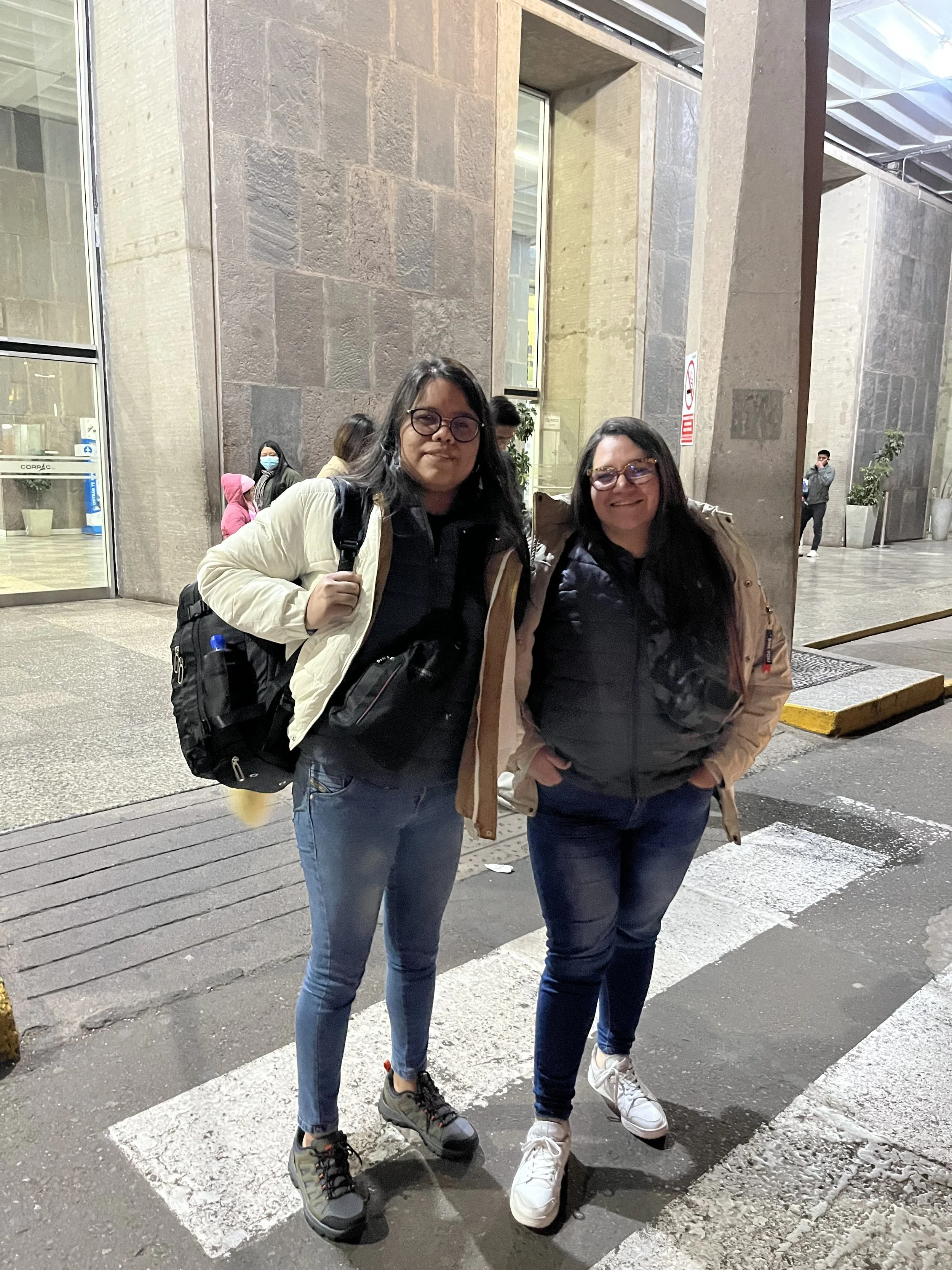 Dos mujeres de pie en la acera frente a un edificio de piedra, una lleva mochila y ambas usan gafas, ropa de abrigo y zapatos deportivos.