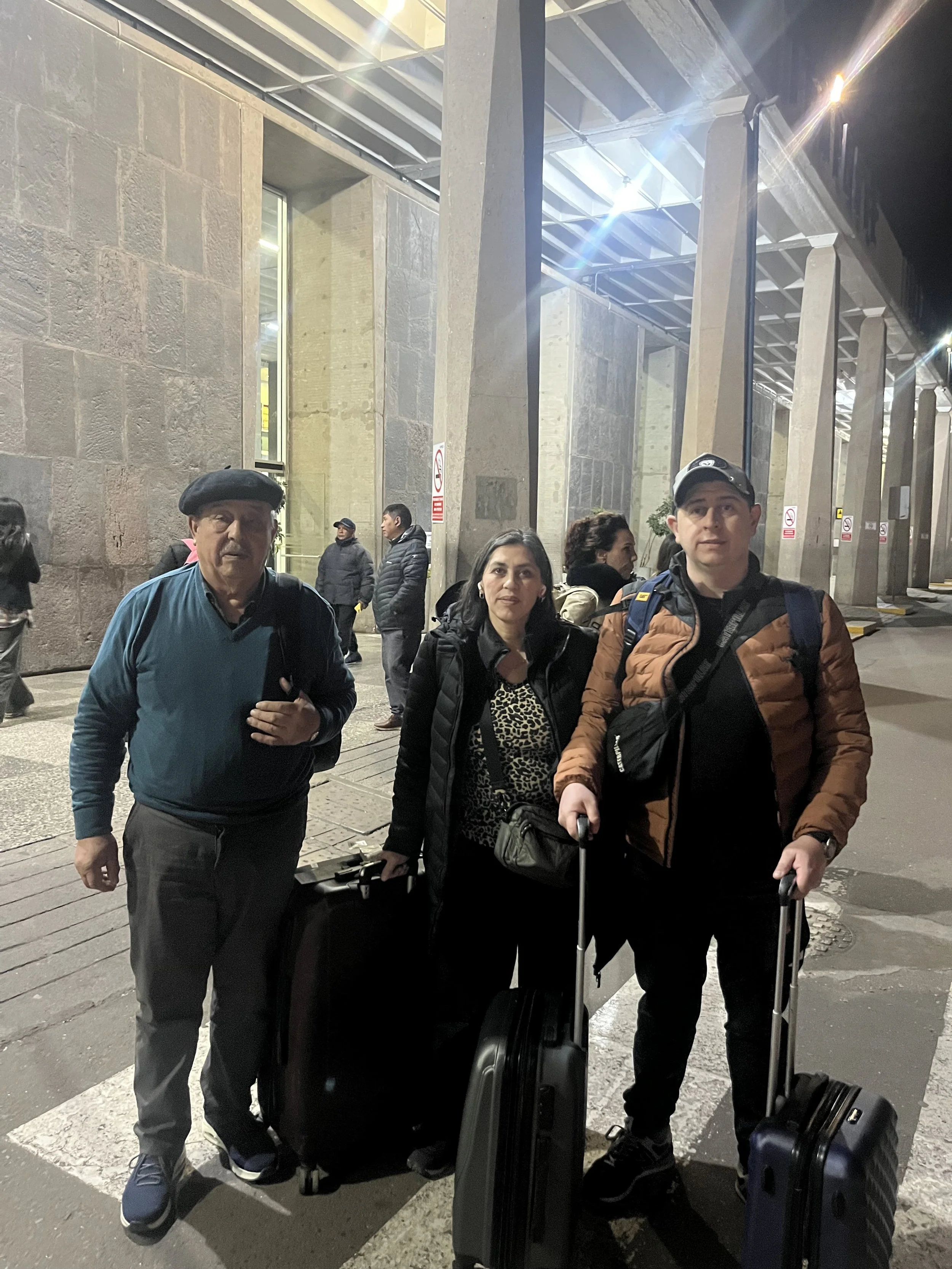 Tres personas con equipaje en la entrada de una estación de tren o aeropuerto por la noche.