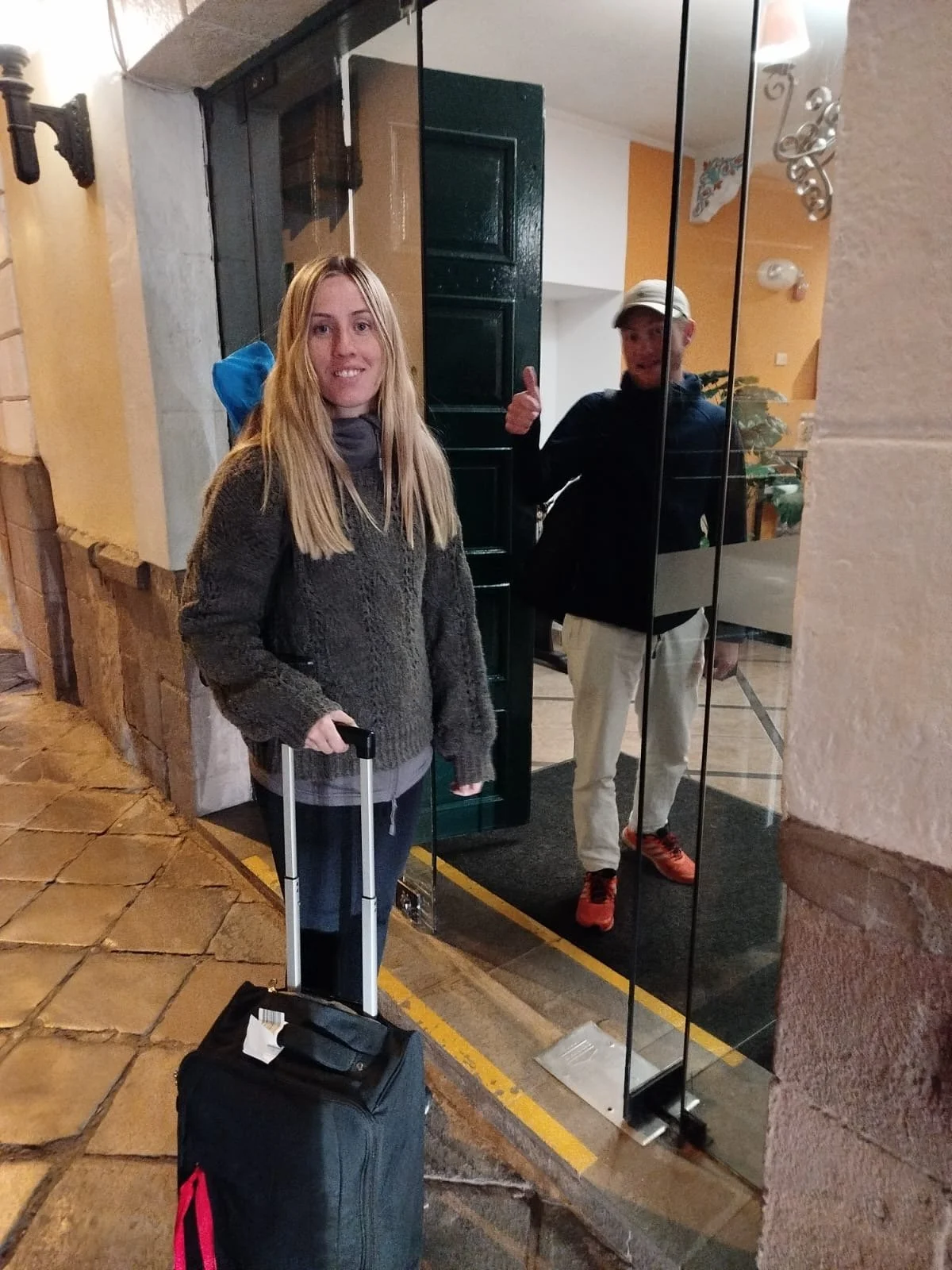 Una mujer con equipaje de mano frente a un espejo en la entrada de un edificio, acompañado de un hombre que da un pulgar arriba en el fondo.