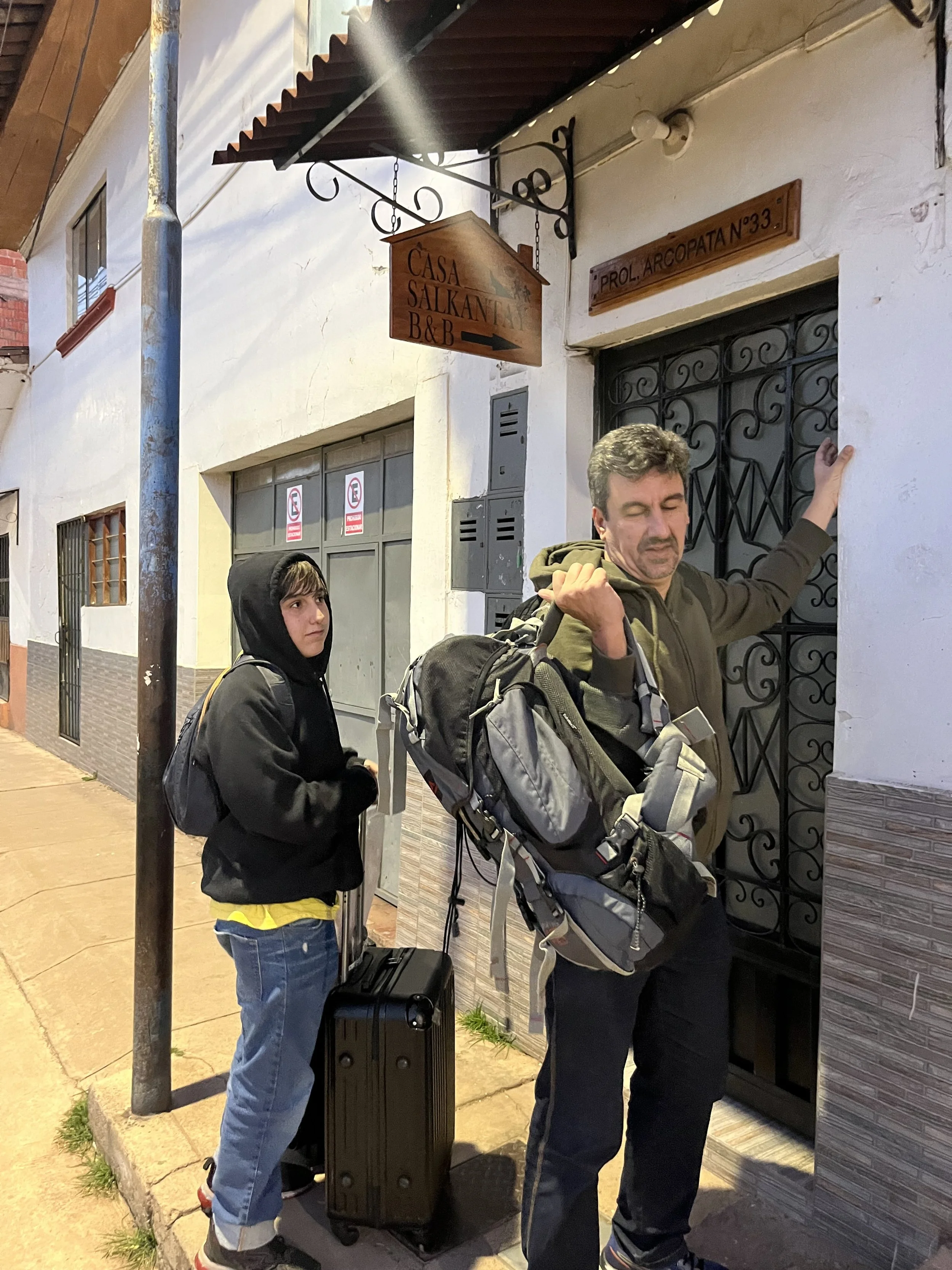 Dos personas con mochilas y equipaje esperan frente a una puerta de entrada a una casa o posada llamada Casa Salkantay, con un cartel que indica dirección y un entorno urbano nocturno.