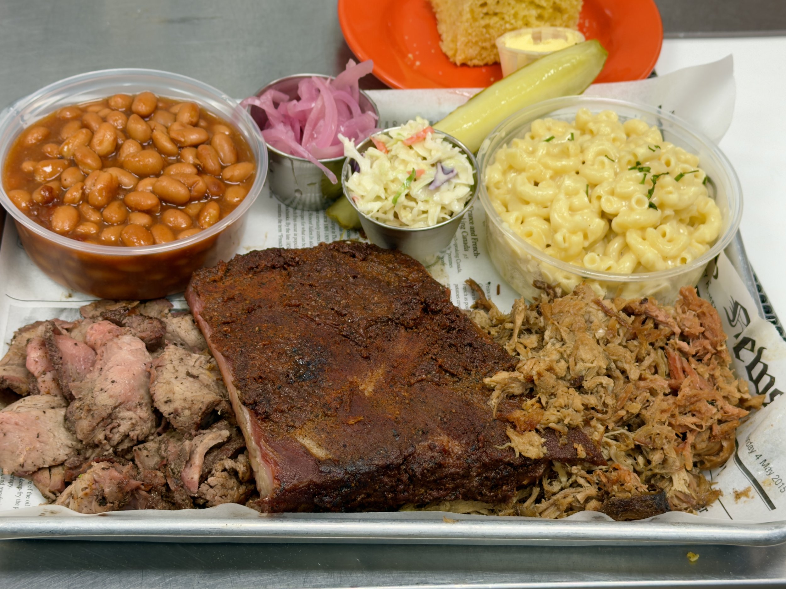 Tray with baked beans, sliced barbecue pork, brisket with sauce, shredded barbecue meat, macaroni and cheese, pickles, coleslaw, sliced onion, sliced pickles, corn muffin with butter, and pickle spear.