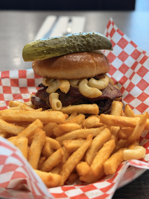 A cheeseburger with a pickle, bun, macaroni salad, beef patty, and French fries on red-checkered paper.