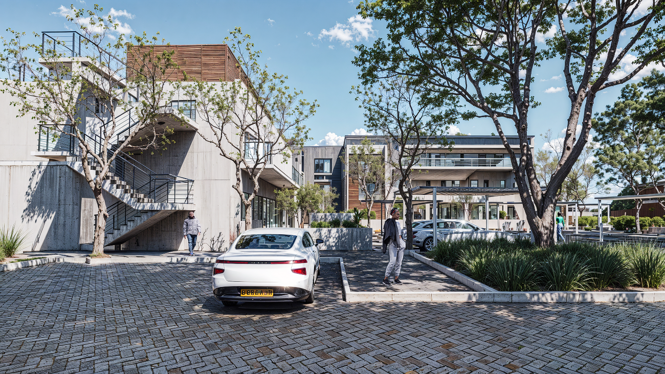 A modern residential apartment complex with trees, parked cars, and people walking in the parking lot on a sunny day.