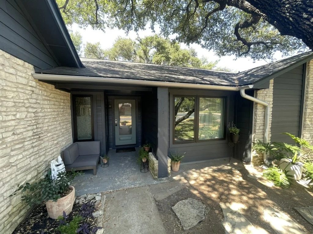 Front entrance of a house with a gray door, black framing, and large windows, surrounded by plants and a stone patio.