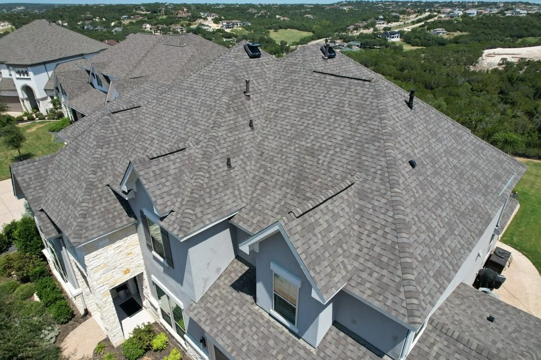 Aerial view of a large residential house with gray shingle roof and multiple gables, surrounded by green landscape.