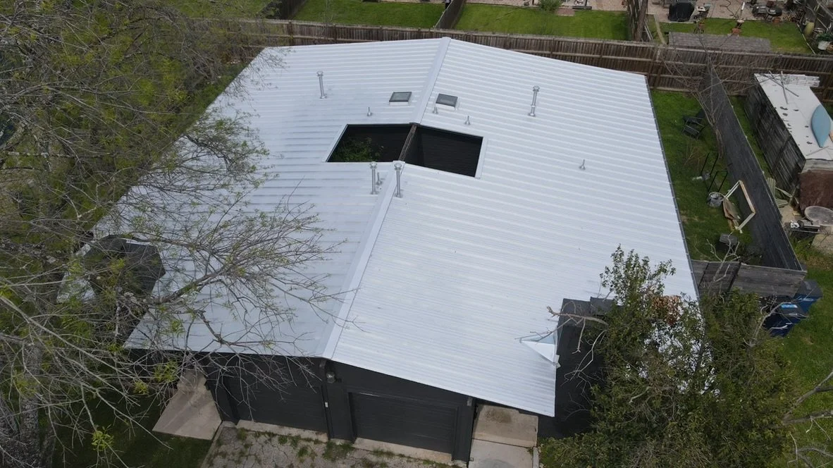 Aerial view of a house with a silver metal roof, surrounded by trees and a wooden fence, with a driveway and some outdoor structures nearby.