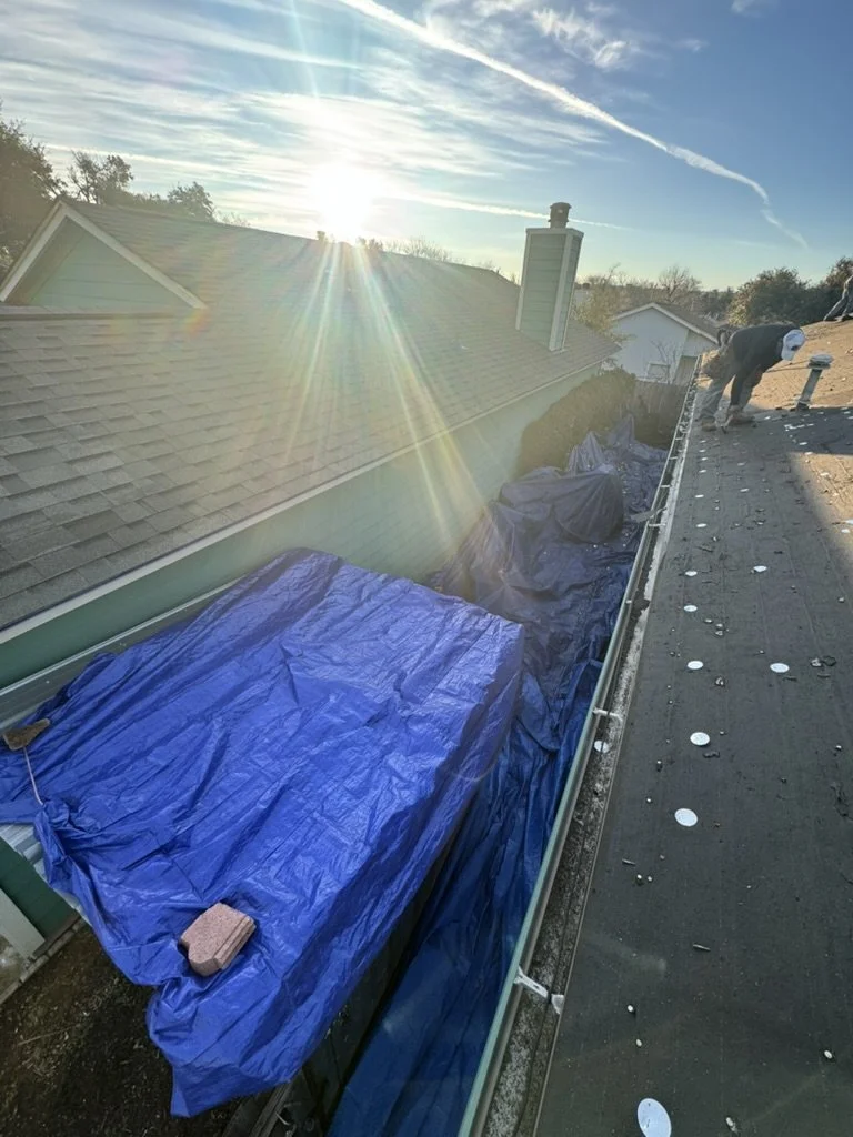 A roof under construction with a worker installing shingles; blue tarp covers part of the roof, and the sun is shining brightly in a clear sky.
