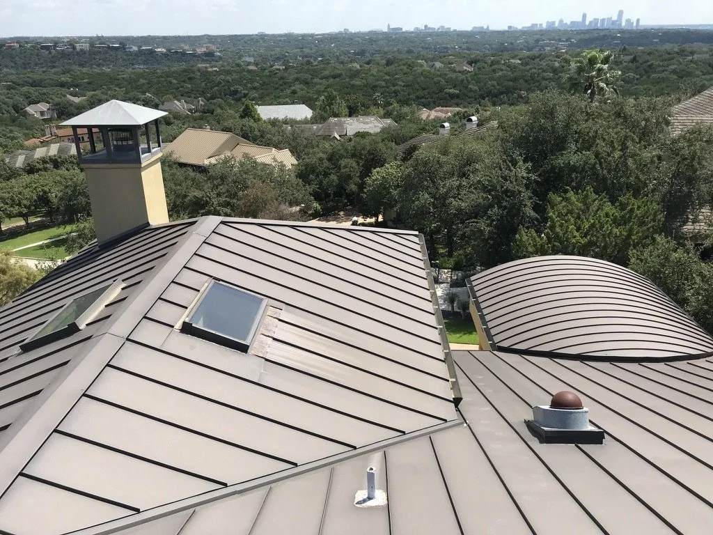 A metal roof with skylights, a roof vent, and a chimney, overlooking a residential neighborhood with greenery and a city skyline in the distance.