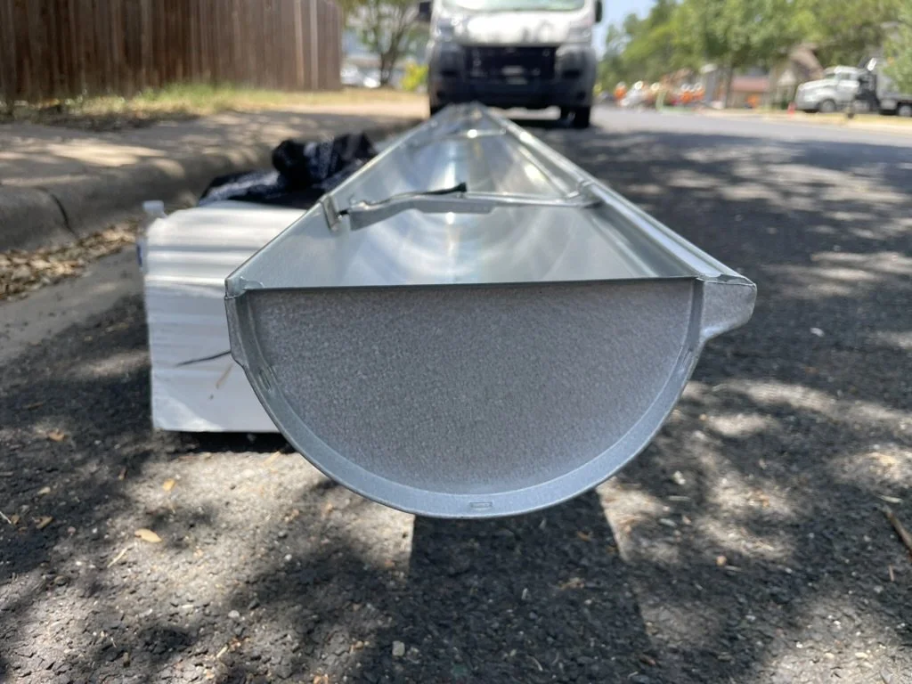 View of a metallic rectangular object on the ground in a neighborhood street, with a semi-circular metal component pointing towards the camera, cars and trees in the background.