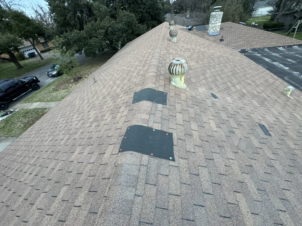 Aerial view of a house roof with multiple roof vents and patches, neighboring trees, cars parked on the street, and partly visible neighboring houses.