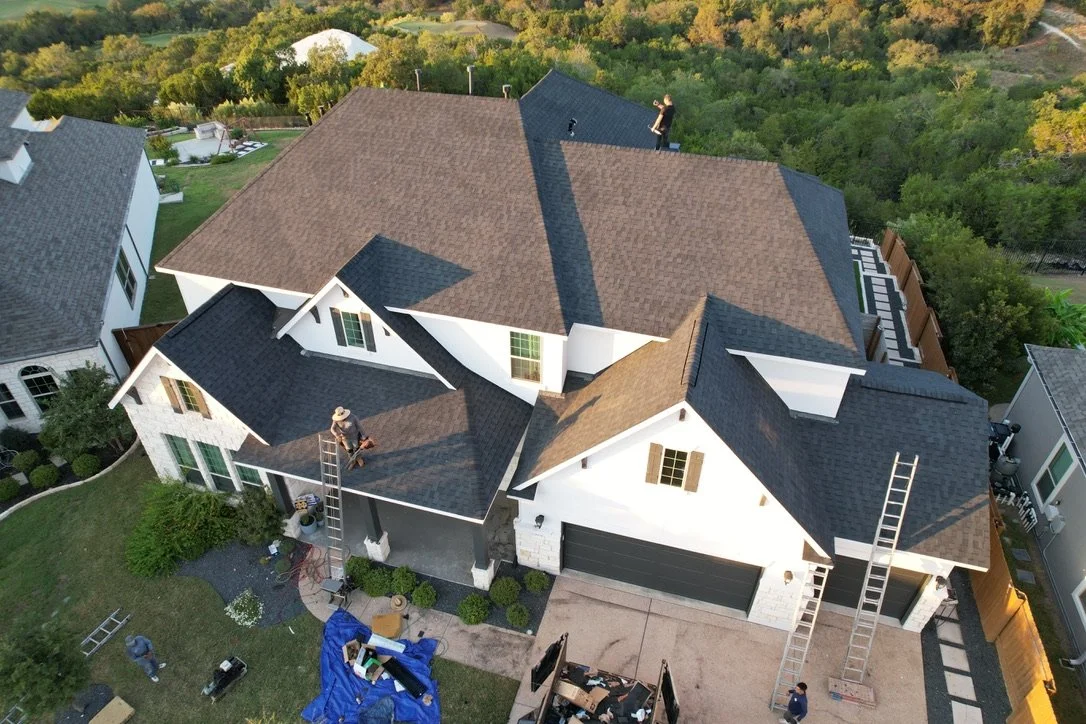 An aerial view of a house with workers on the roof installing shingles, surrounded by other houses and greenery.