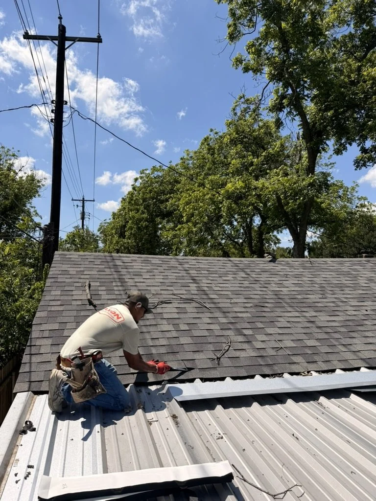 A worker installing or repairing a metal roof on a house, with trees and power lines in the background.