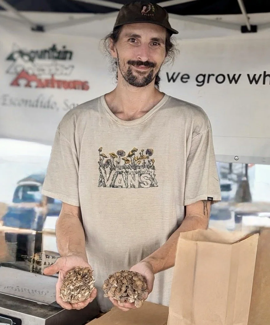 A man with a beard and mustache wearing a black cap and beige Vans t-shirt holding two large mushrooms at a market stand. The background features a banner with partially visible text and cars in the distance.