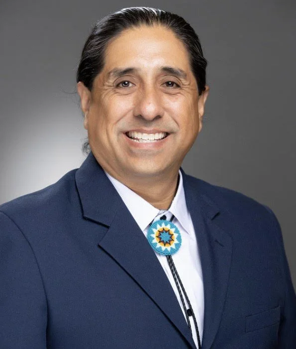 A man with dark hair in a black suit and white shirt, smiling, wearing a decorative bolo tie with a circular blue and yellow design, against a gray background.