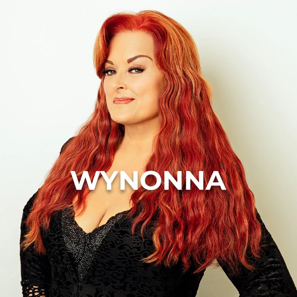 A woman with long, wavy red hair, wearing a black dress with a textured pattern, facing the camera with a confident expression. The word "WYNONNA" is overlaid on her chest.