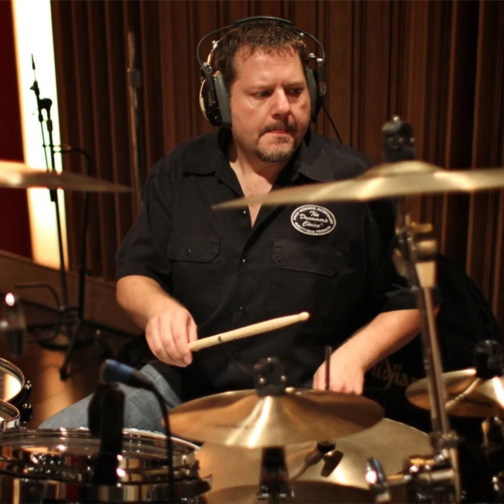 A man with short brown hair, wearing headphones and a black button-up shirt, is playing the drums in a music studio.