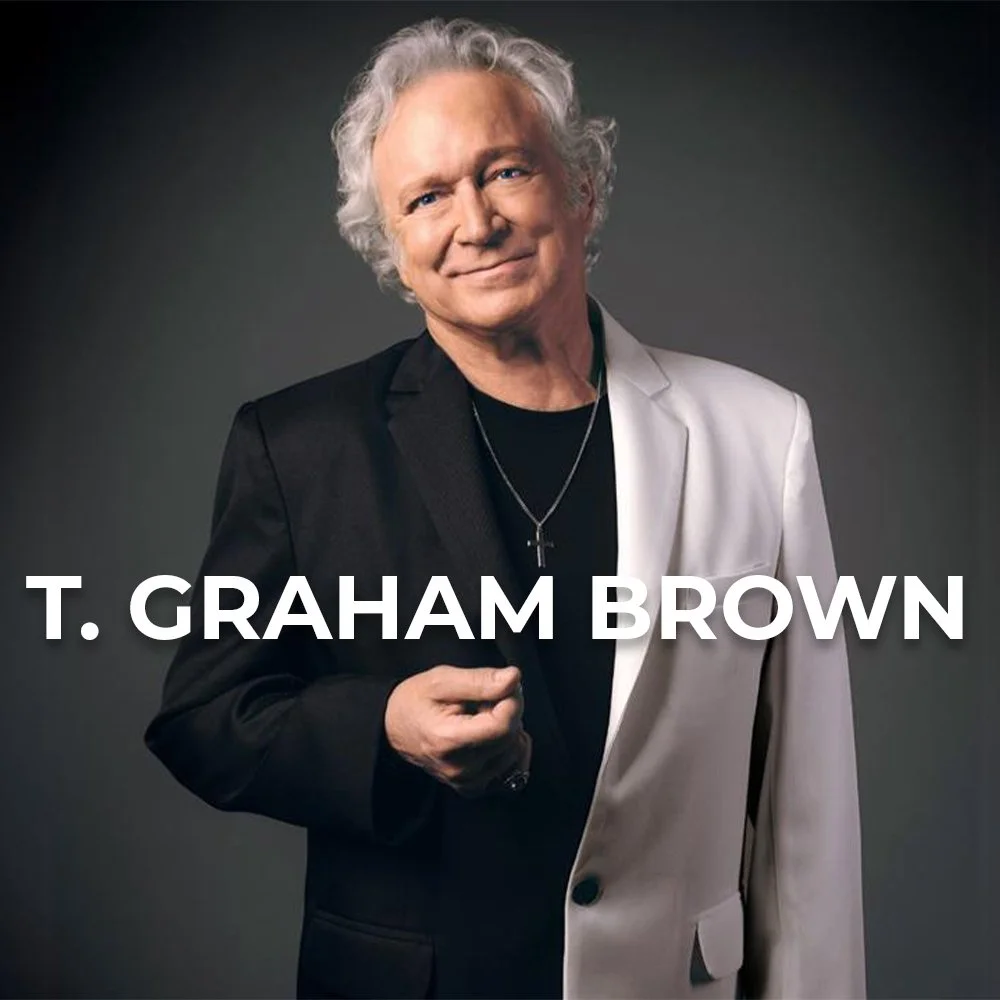 Portrait of T. Graham Brown, a man with white curly hair, wearing a white blazer on one side and a black blazer on the other, smiling, with a gray background, and the text 'T. Graham Brown' overlaid.