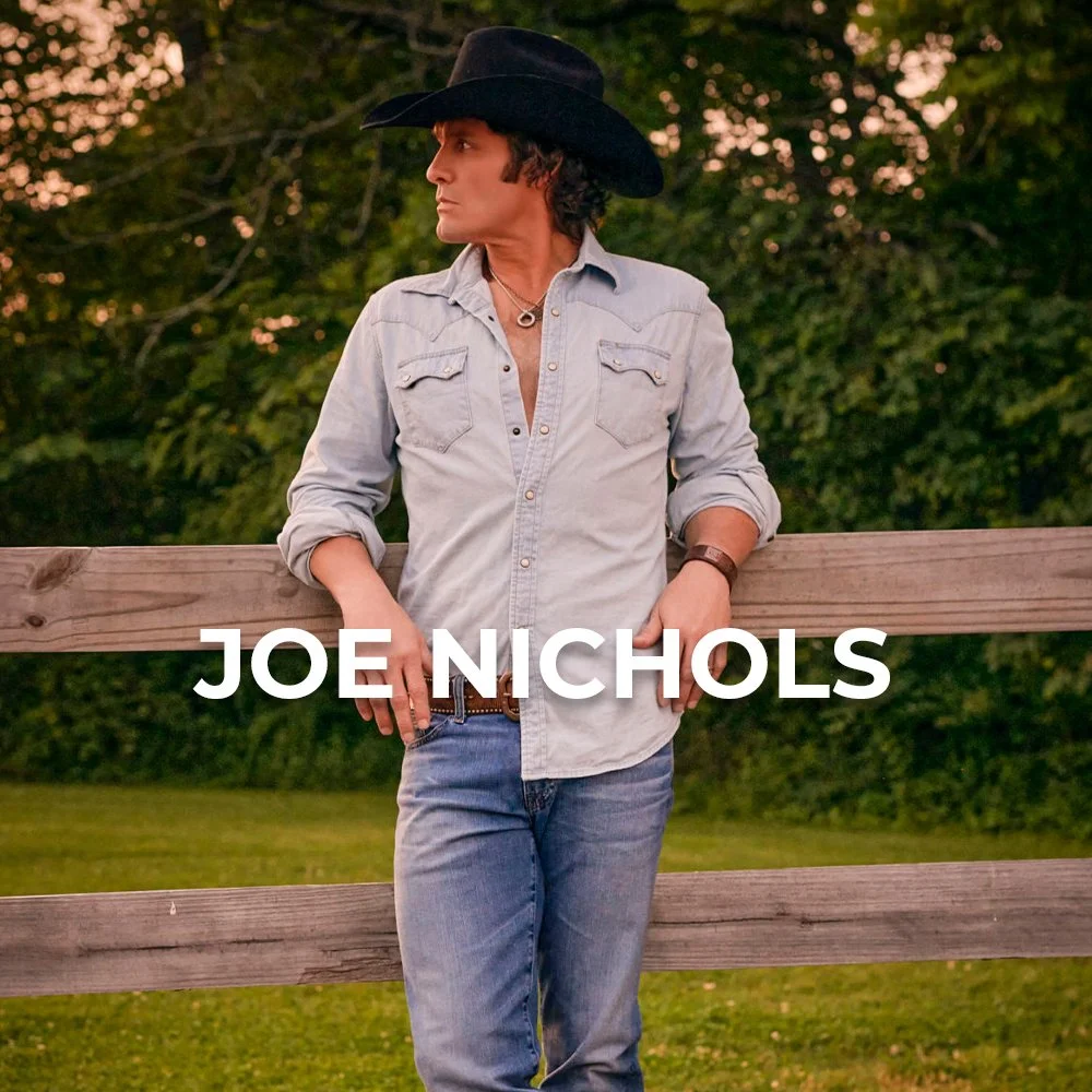 A person wearing a black cowboy hat, denim shirt, and jeans leaning on a wooden fence outdoors with trees and greenery in the background.