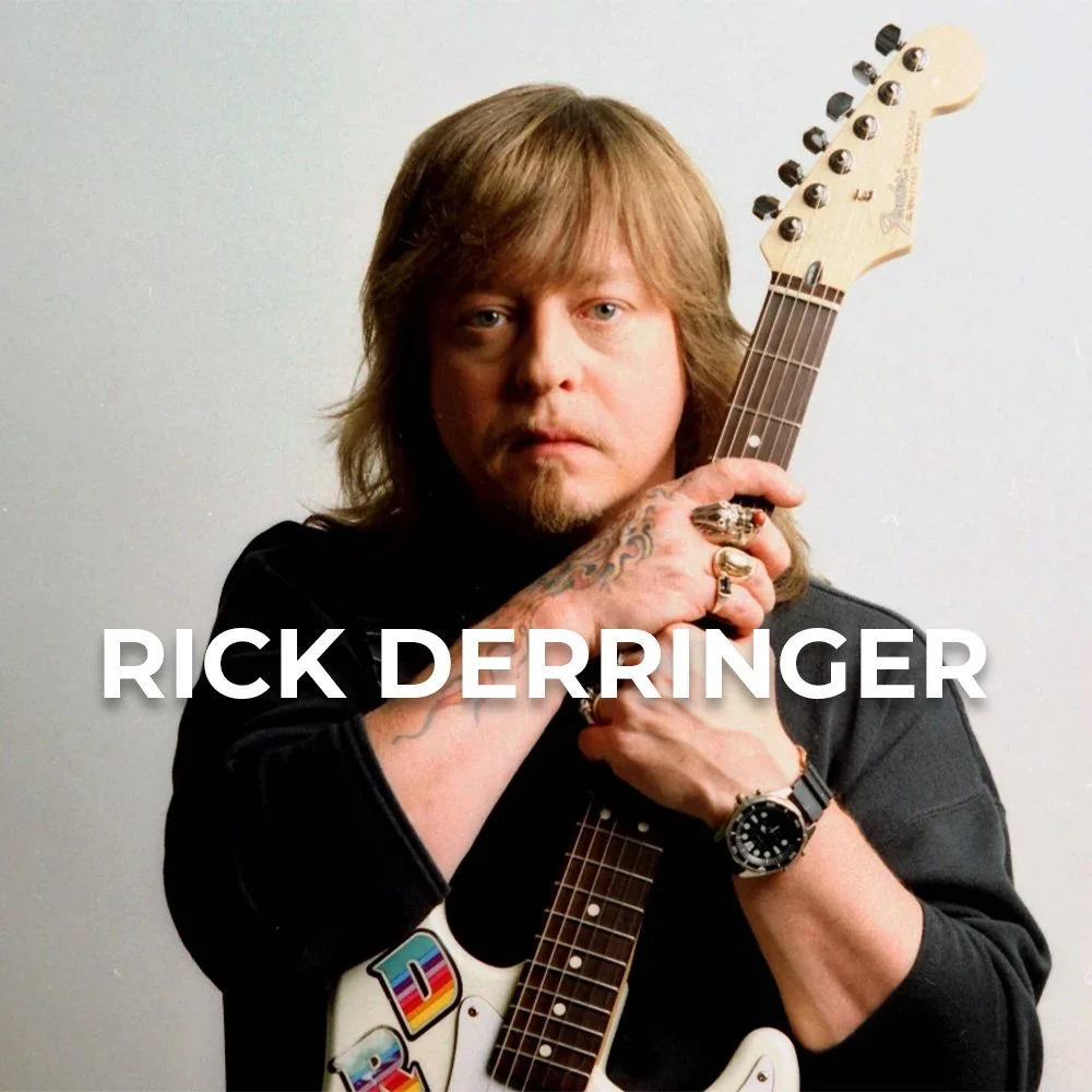 A man with long, light brown hair, tattoos on his hand, holding an electric guitar with the neck resting on his face, standing against a light background. The text 'Rick Derringer' is overlaid on the image.