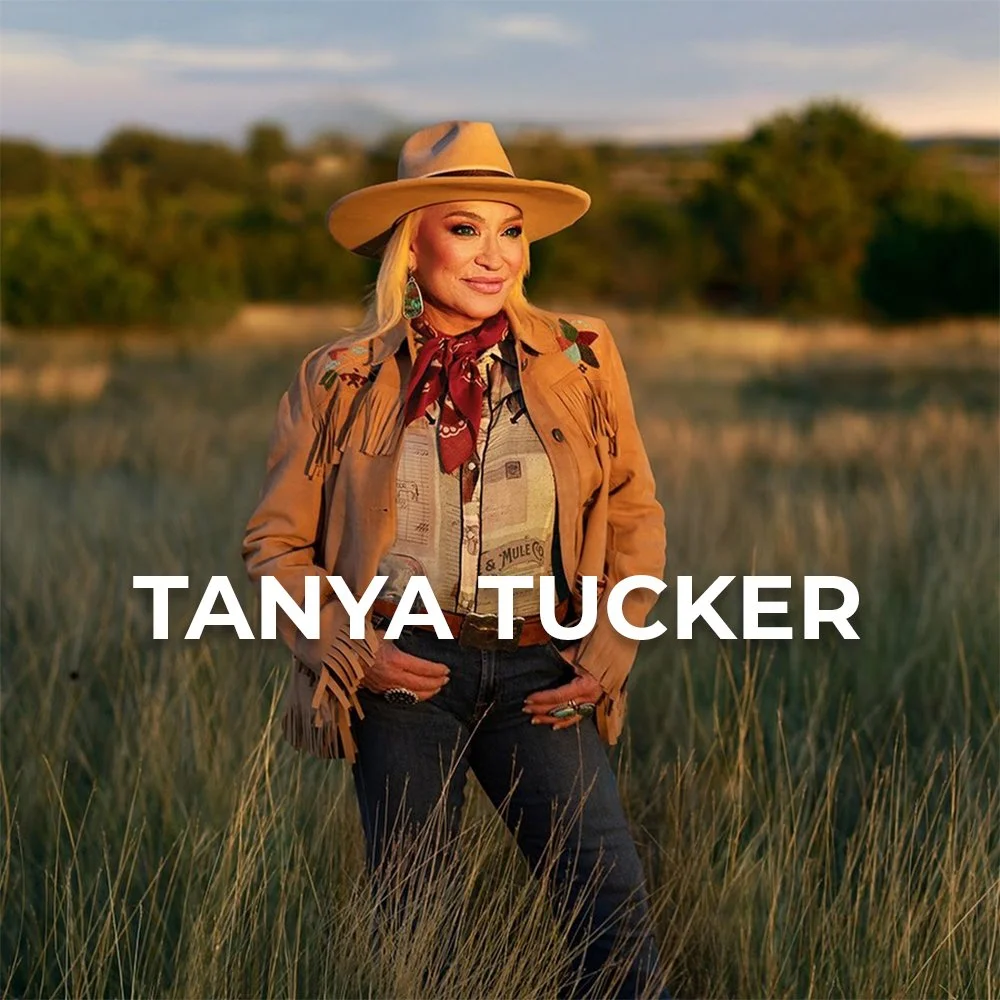 Tanya Tucker posing outdoors in a field with tall grass, wearing a cowboy hat, a fringed jacket, a bandana around her neck, and dark jeans, with a scenic rural background at sunset.