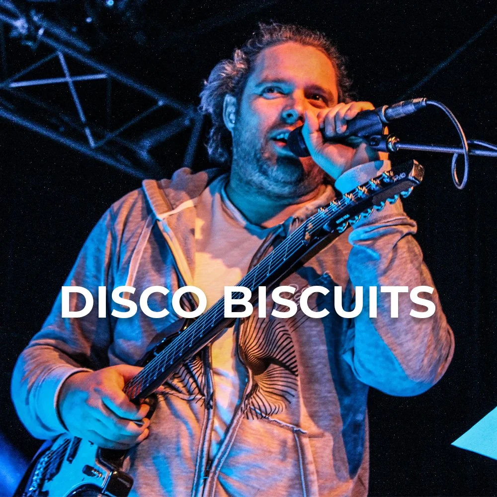 A man singing into a microphone while playing an electric guitar, under stage lighting with a dark background and metal truss.
