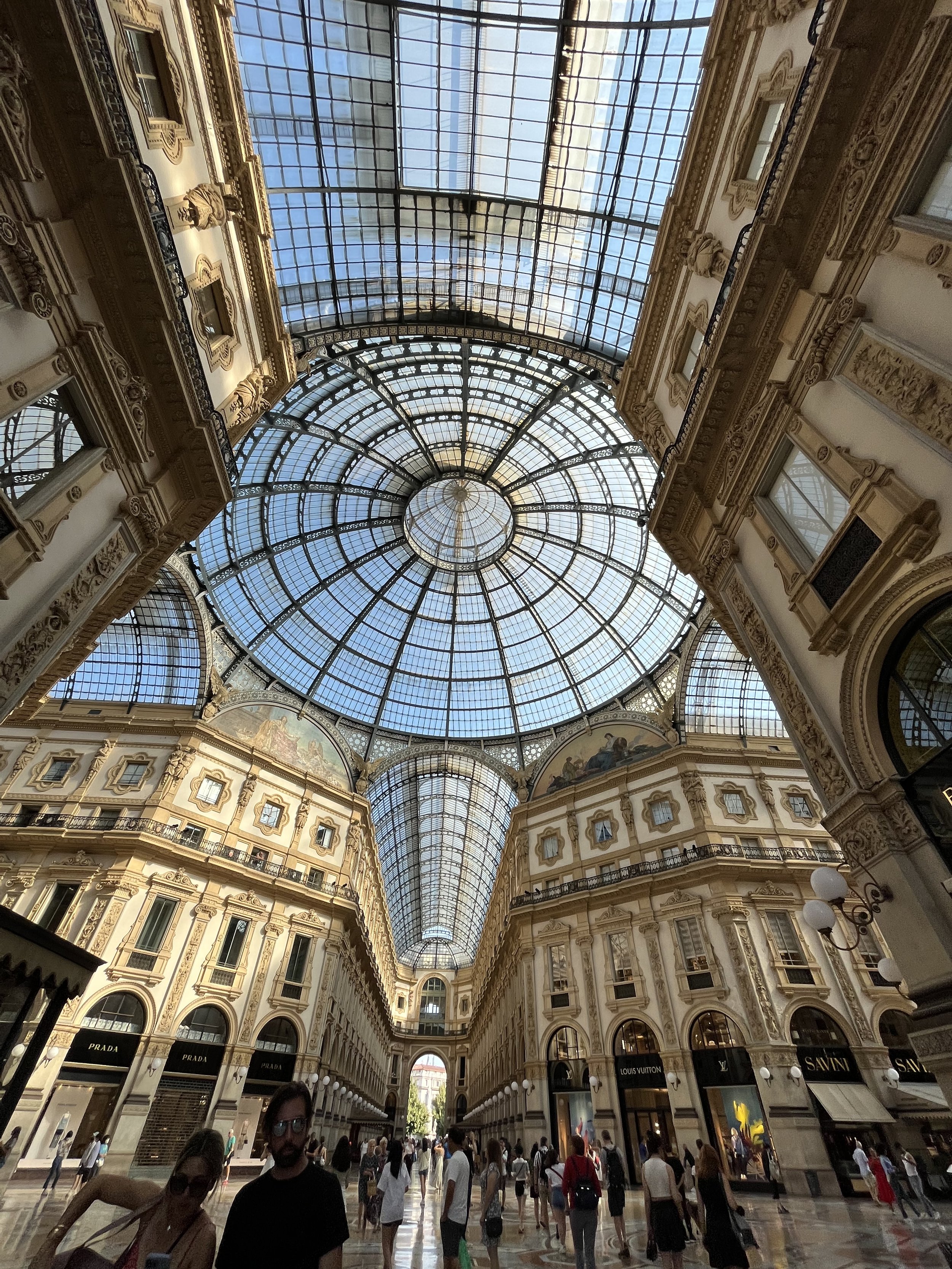 Interno di un centro commerciale o galleria con grande soffitto in vetro e decorazioni in stile classico, molte persone camminano lungo il passaggio centrale.
