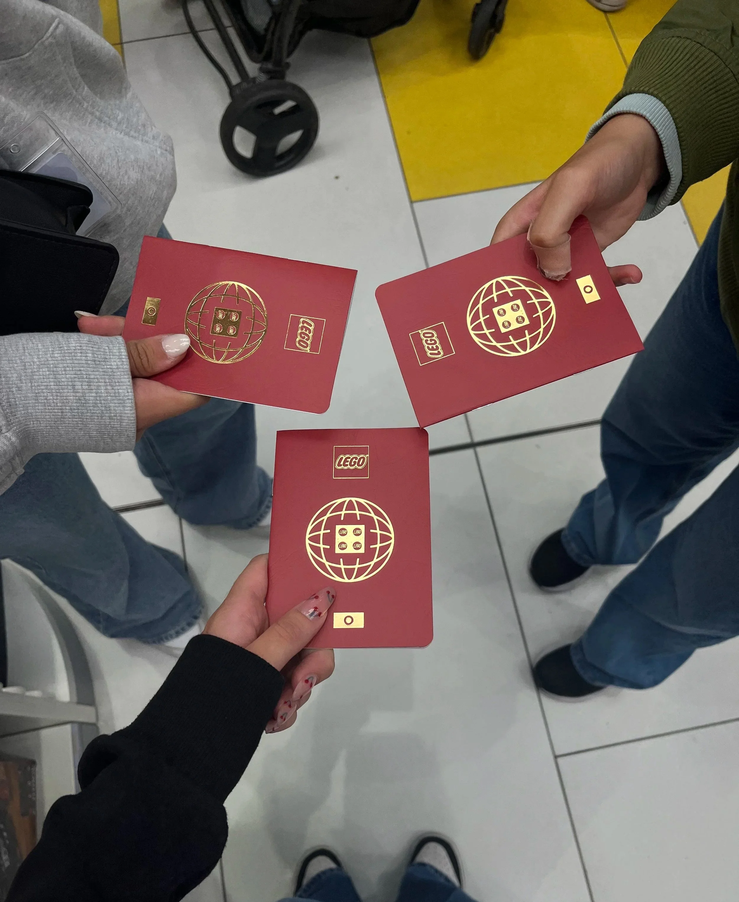 Tre persone tengono in mano dei passaporti rossi con un logo dorato di LEGO e un globo stilizzato, in un ambiente con pavimenti bianchi e gialli e in presenza di altri oggetti come una sedia e una carrozzina.