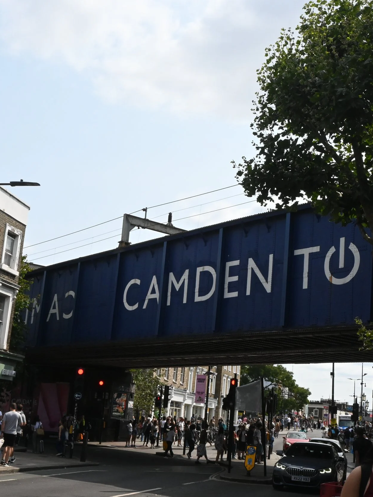 Immagine di un incrocio trafficato con molte persone che attraversano, automobili e un grande cartellone blu sopra la strada con la scritta 'CAMDEN T'. Ci troviamo a Camden Market a Londra.