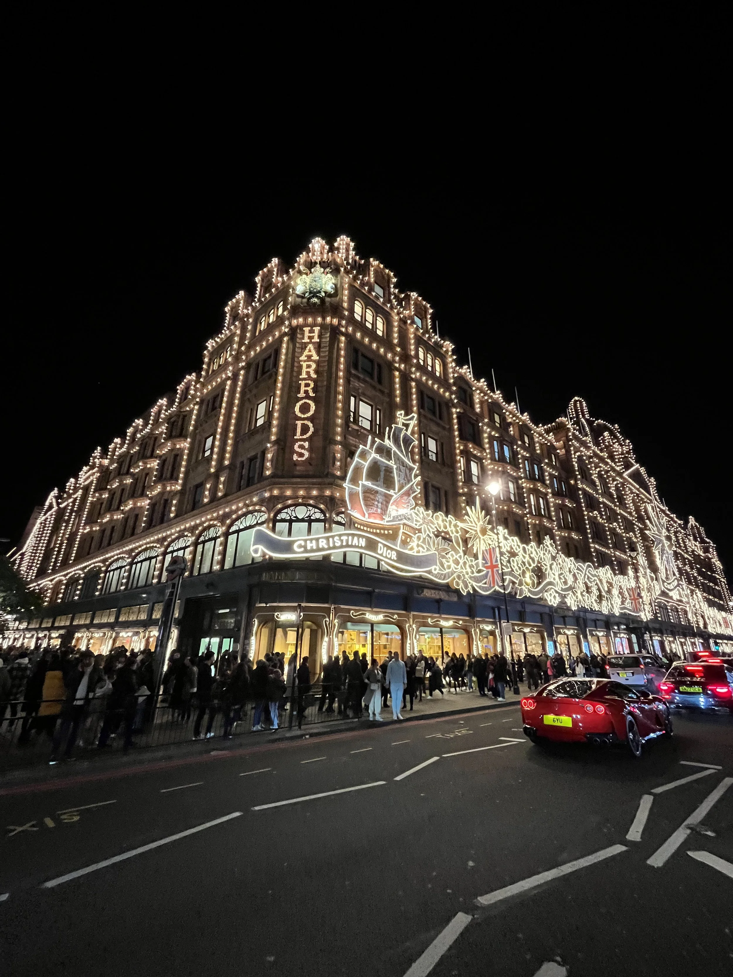 Edificio addobbato con luci natalizie, segnato con il nome Harrods, in una notte con molte persone e auto sulla strada. Harrods a Londra.