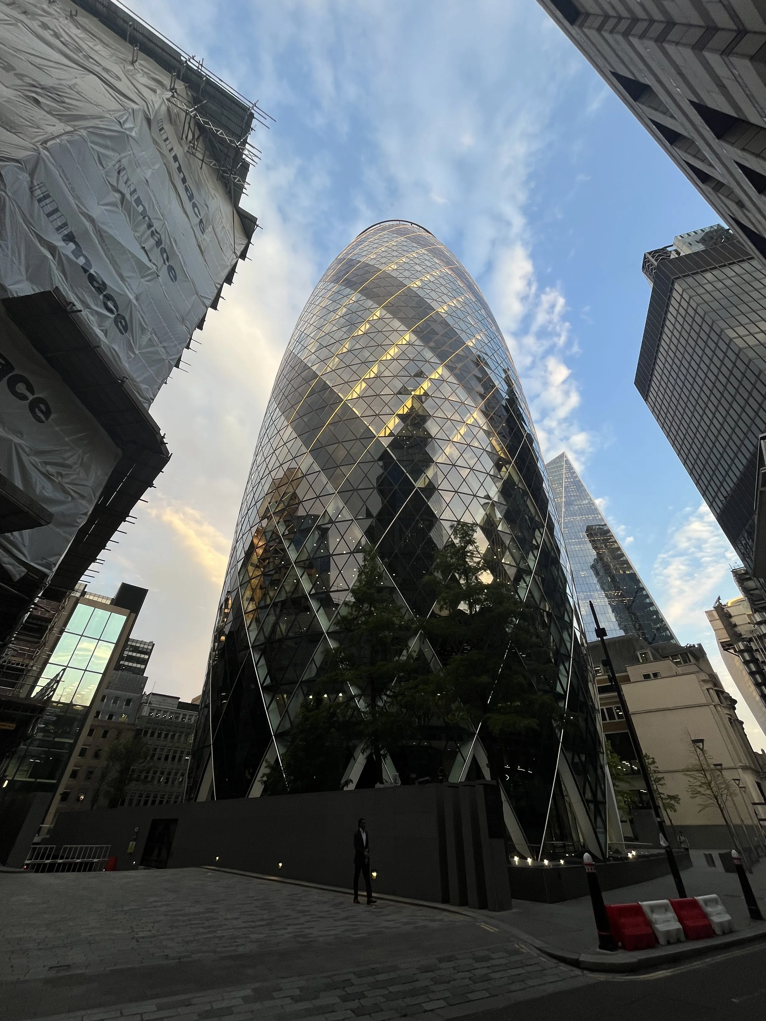 Edificio moderno con facciata di vetro e forma ovale, circondato da altri grattacieli in un'area urbana, con il cielo azzurro e nuvole. the gherkin, il cetriolo di Londra.