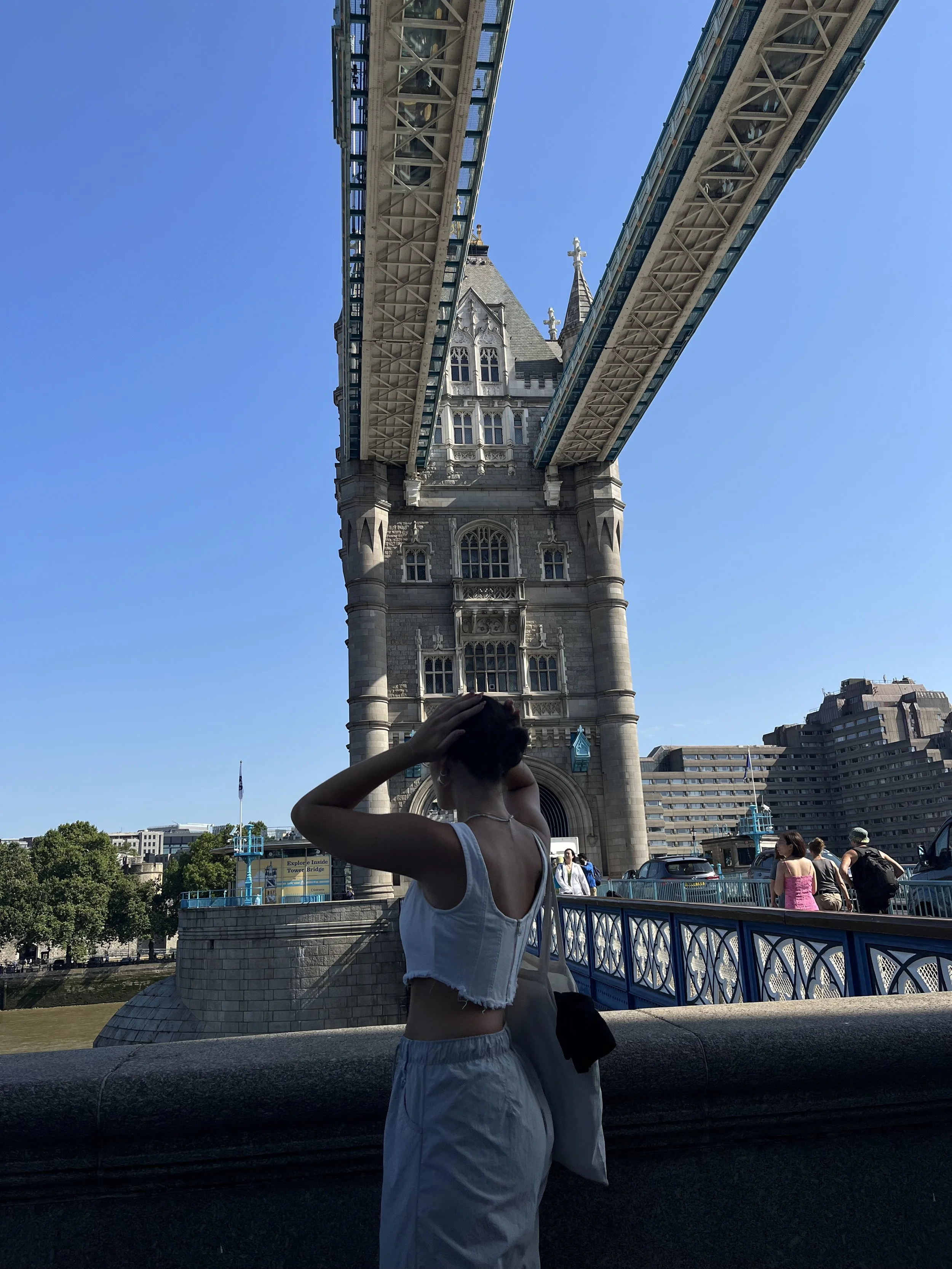 Una donna vestita con top bianco e pantaloni bianchi si trova sotto il Tower Bridge di Londra.
