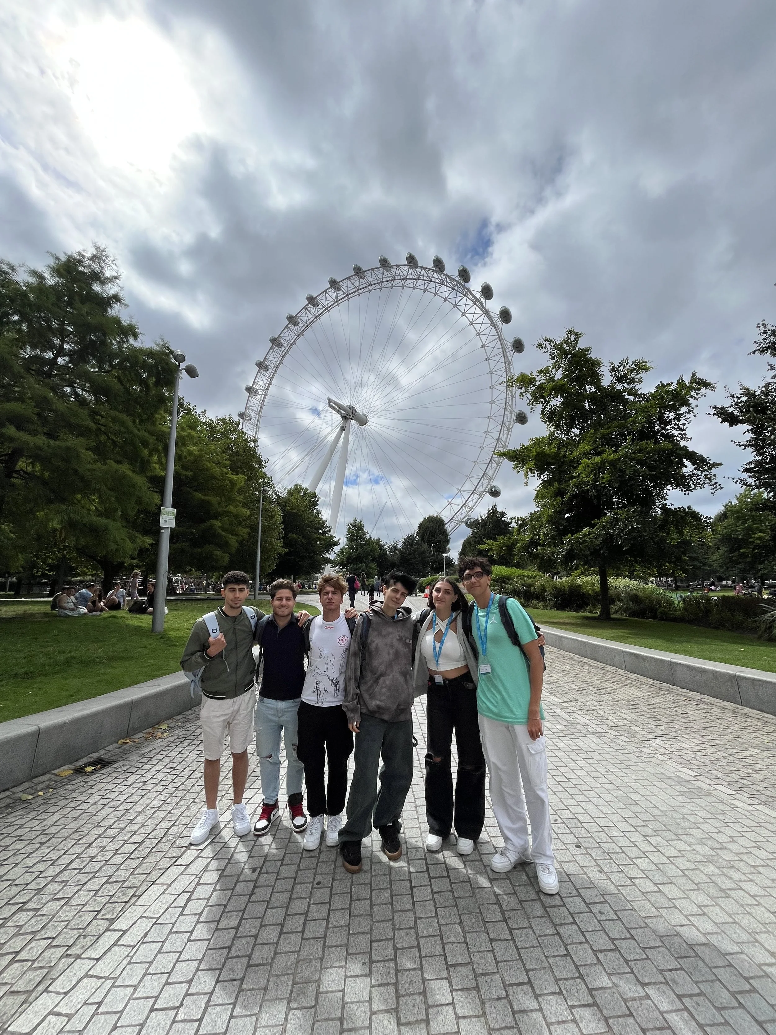 Un gruppo di sei giovani con zaini si trova in piedi su un percorso lastricato, con la ruota panoramica di Londra sullo sfondo e alberi intorno, sotto un cielo nuvoloso.