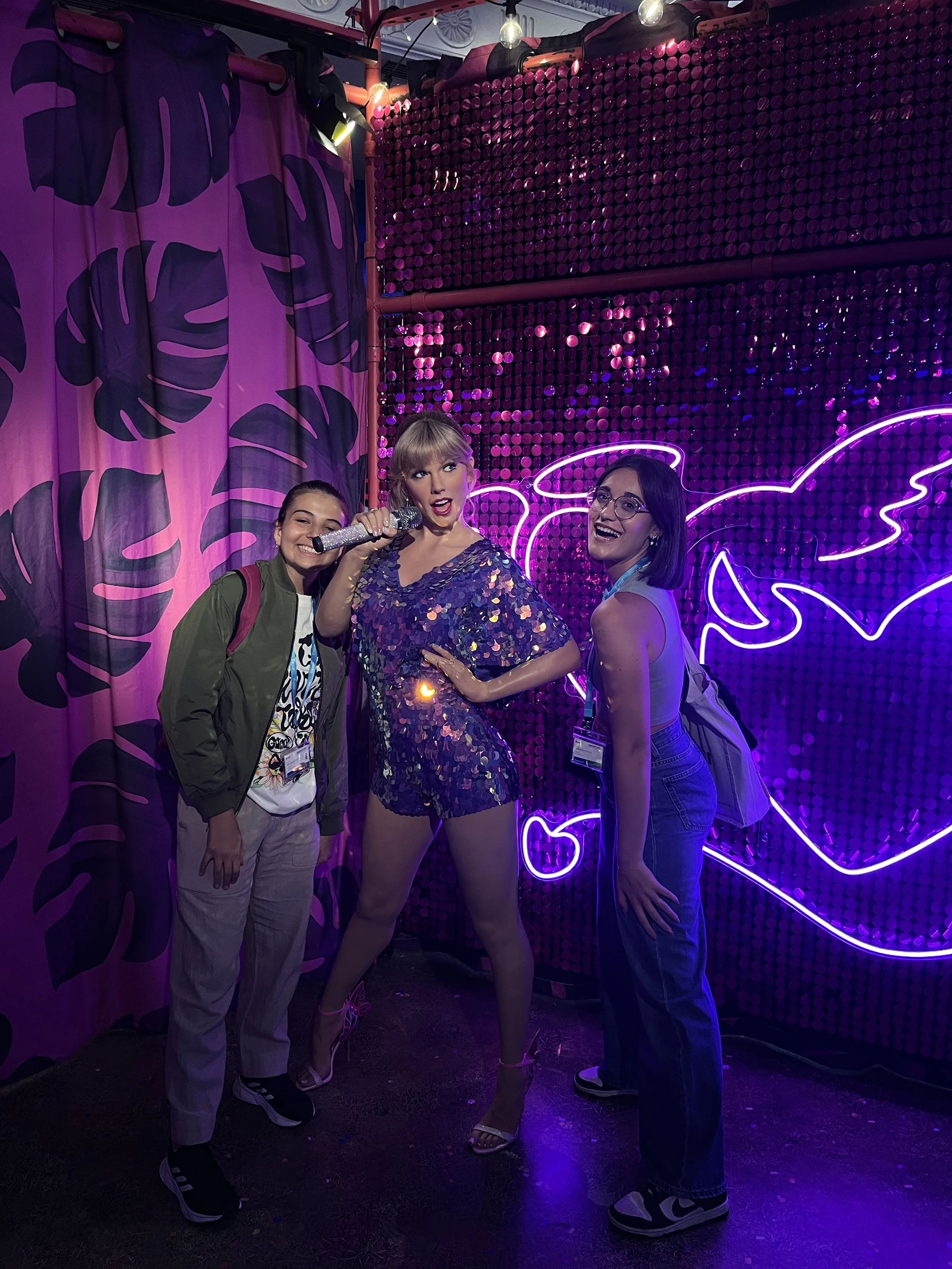 Tre donne sorridenti in abiti colorati, una con microfono, davanti a uno sfondo con luci e decorazioni neon. con taylor swift al Madame Tussauds.