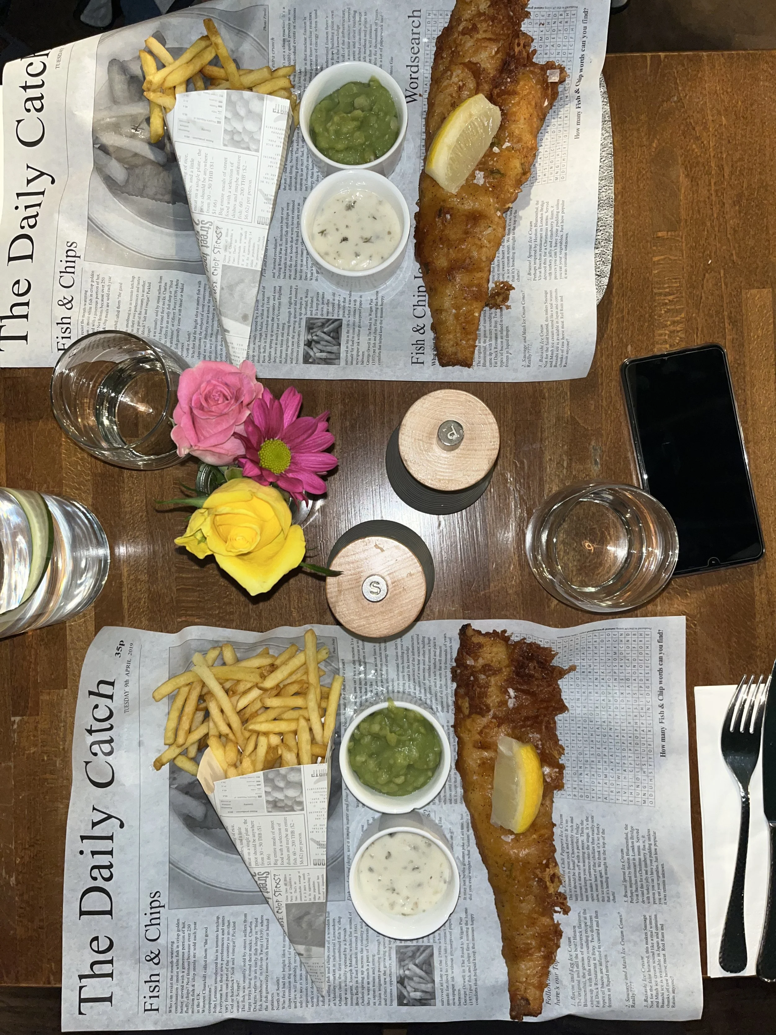 Piatti di merluzzo fritto con insalata di piselli, salsa tartara, patatine fritte, una fetta di limone, e due bicchieri d'acqua su un tavolo con carta da giornale. Fish and chips in un ristorante a Londra
