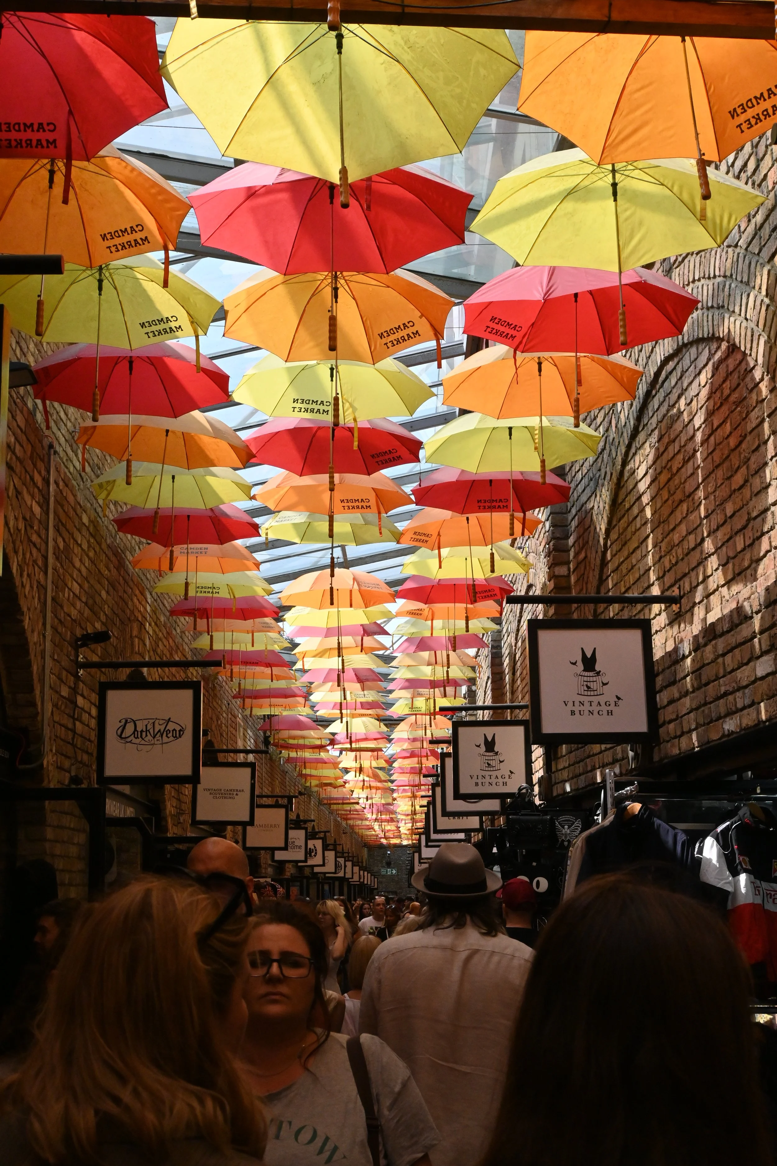 Tetto di ombrelli colorati appesi in un passaggio chiuso da mattoni, con persone che passeggiano. Ci troviamo a Camden Market.