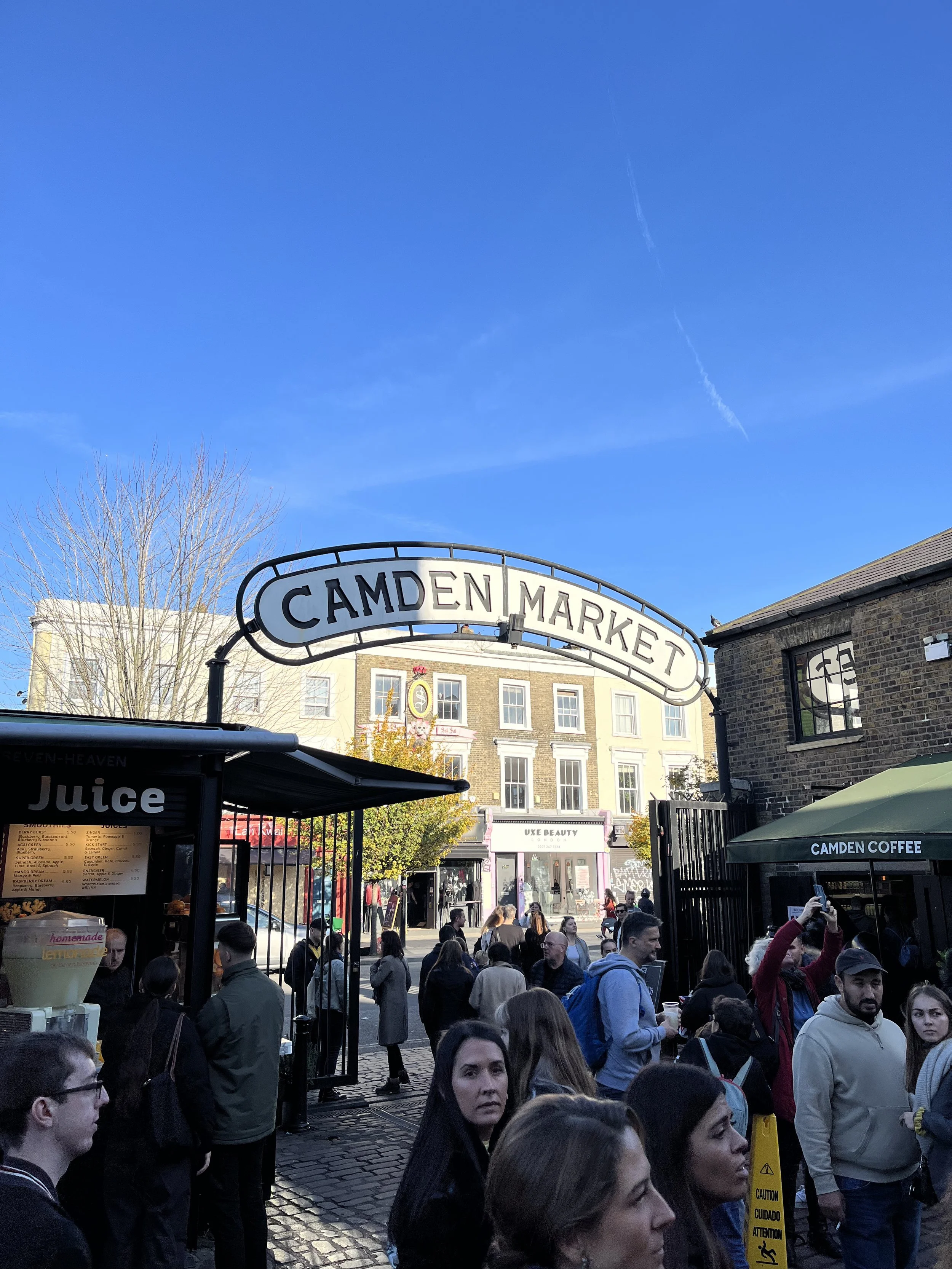 Ingresso a Camden Market con molte persone davanti, negozi e un cielo azzurro sopra.