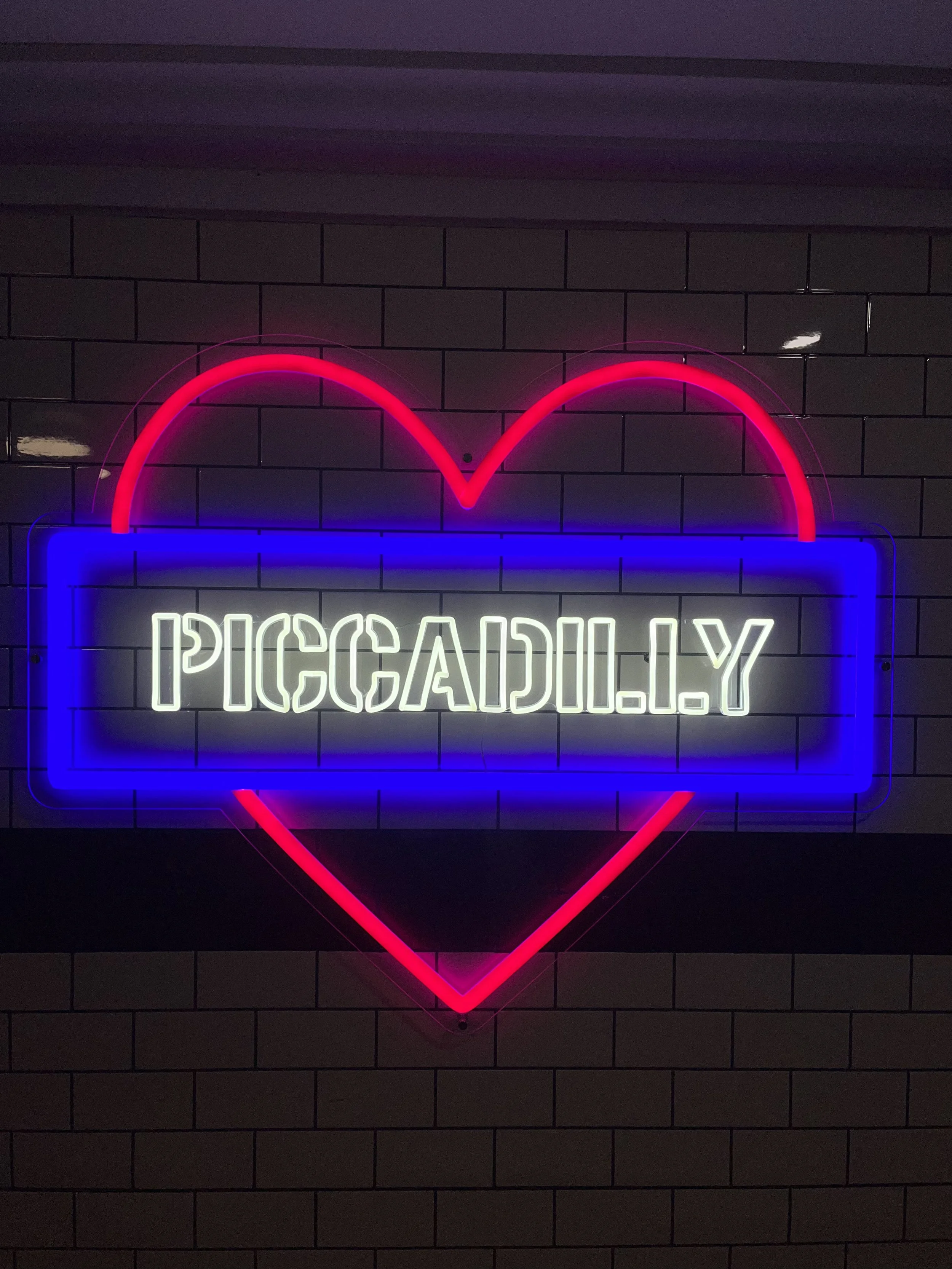 Segnaletica luminosa a forma di cuore con la scritta 'PICCADILLY' in lettere luminose bianche, in un ambiente con pareti in mattoni.