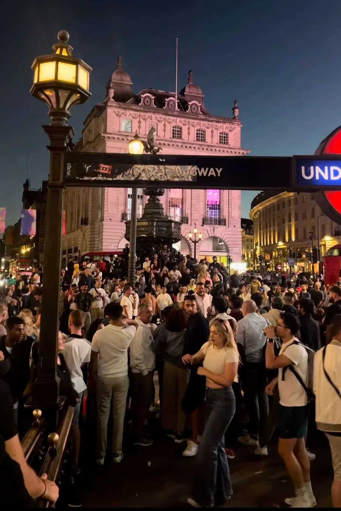 Una grande folla di persone si trova in una zona urbana affollata durante la notte, con un edificio storico illuminato sullo sfondo e un'insegna della metropolitana in primo piano. Ci troviamo a Piccadilly Circus a Londra.