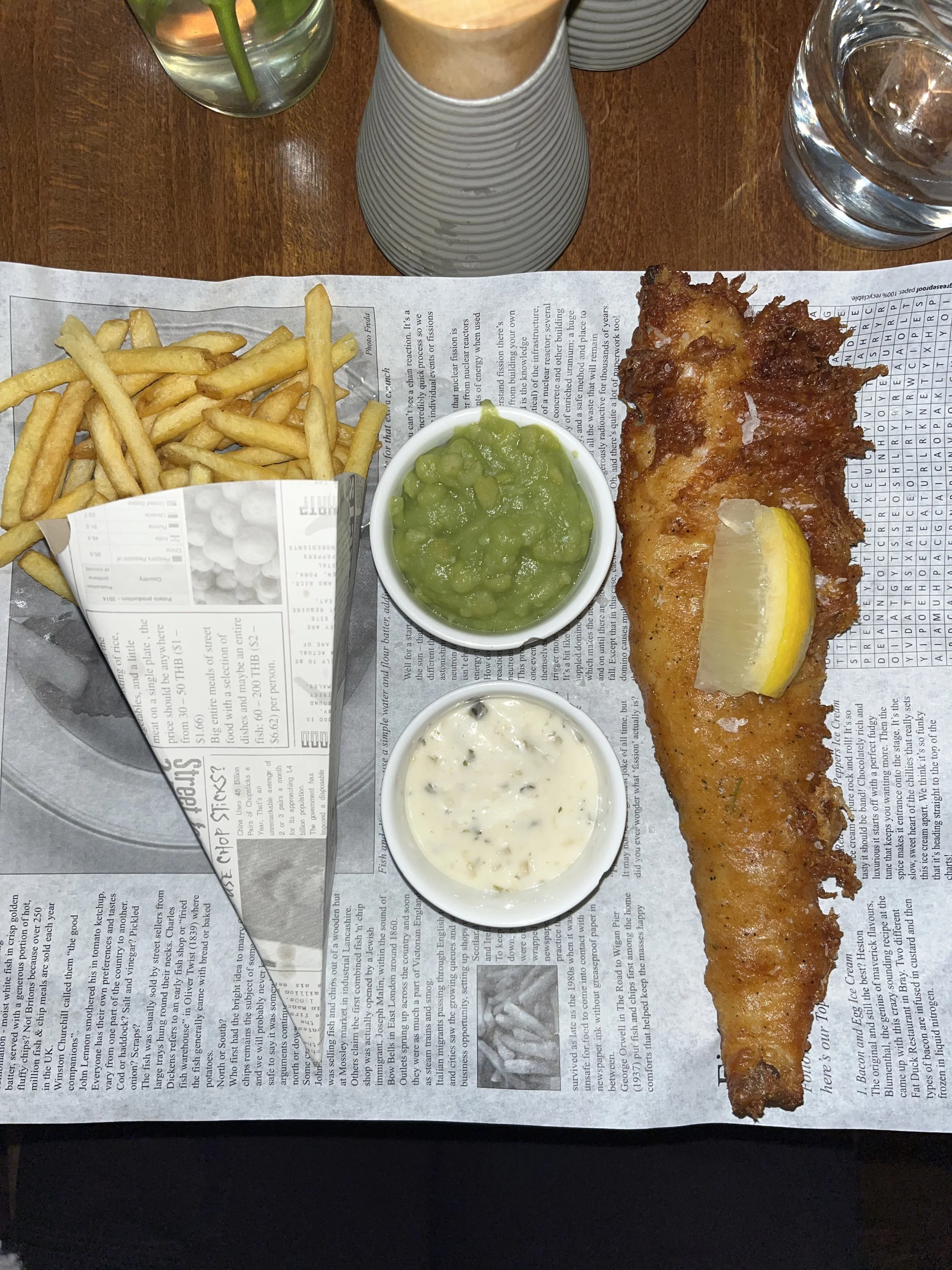Piatti di merluzzo fritto con insalata di piselli, salsa tartara, patatine fritte, una fetta di limone, e due bicchieri d'acqua su un tavolo con carta da giornale. Fish and chips in un ristorante a Londra