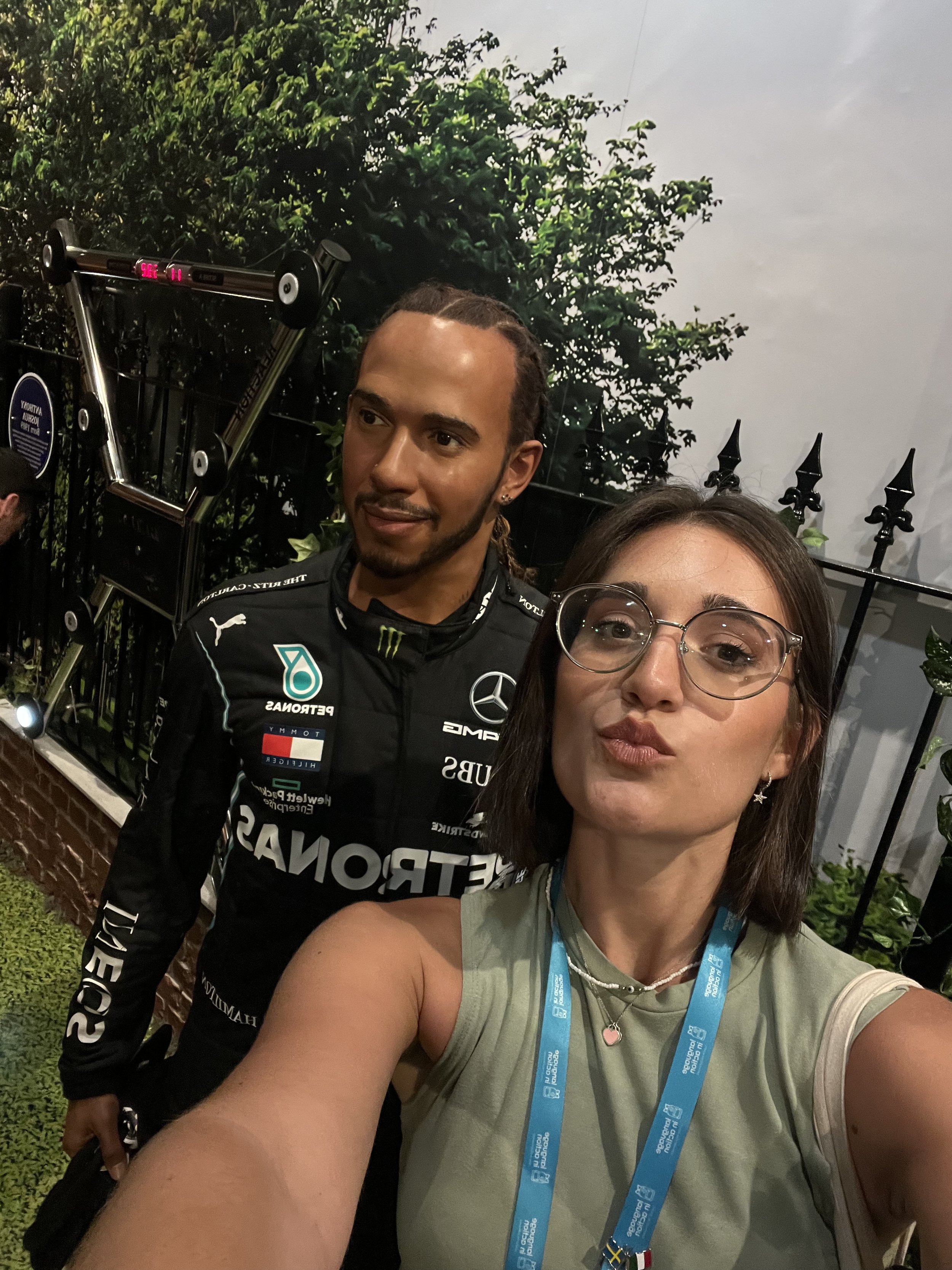Due persone, una donna e un uomo, fanno un selfie insieme. La donna indossa occhiali e una maglietta senza maniche, la donna ha un colletto con un ciondolo a forma di cuore, e una catenina. L'uomo indossa la tuta del team di Formula 1 Mercedes, con v
