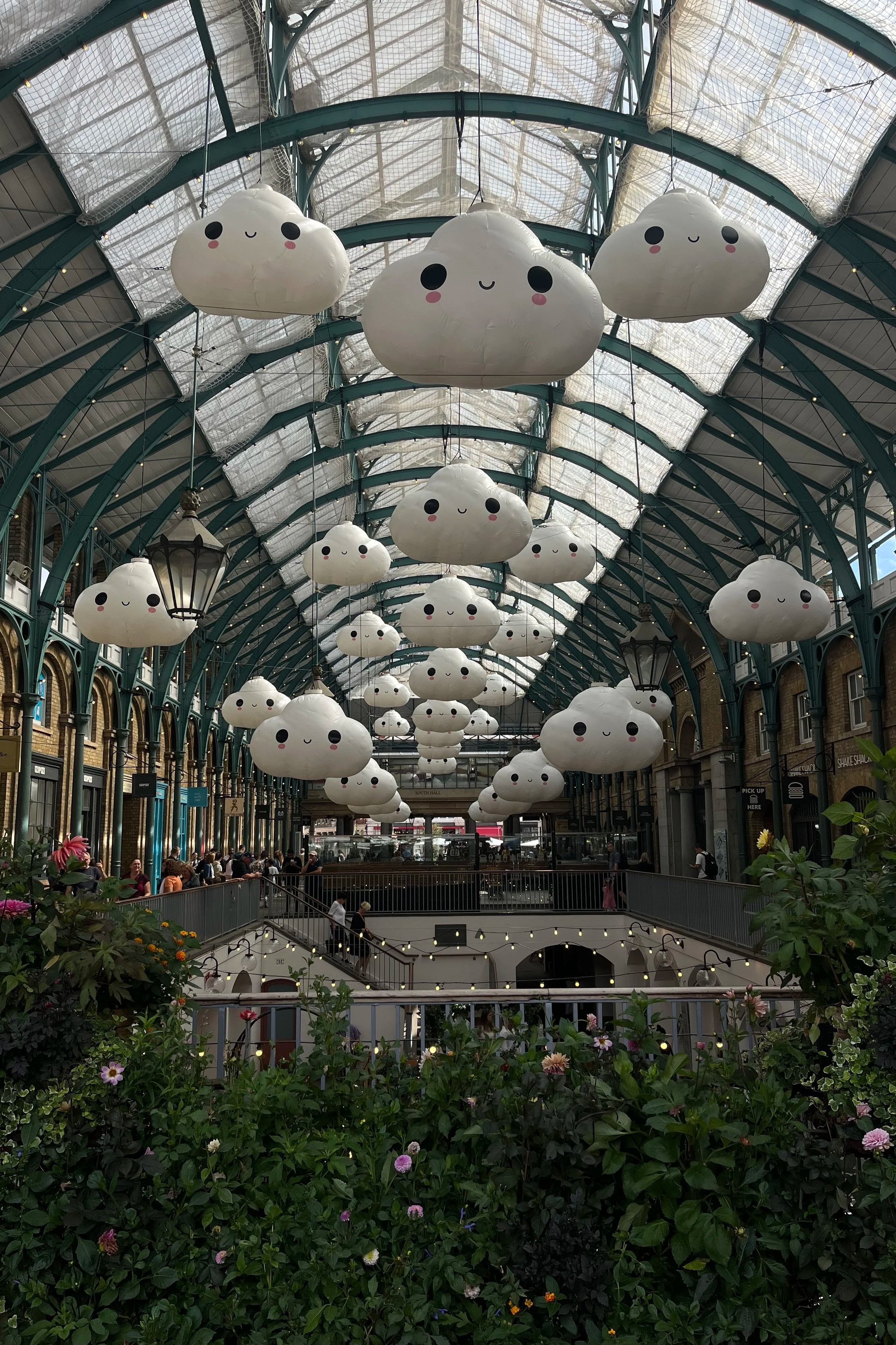 Interno di un mercato o centro commerciale con soffitto di vetro e grandi lanterne a forma di nuvole con faccine sorridenti sospese sul soffitto, decorazioni floreali in primo piano. Covent Garden a Londra