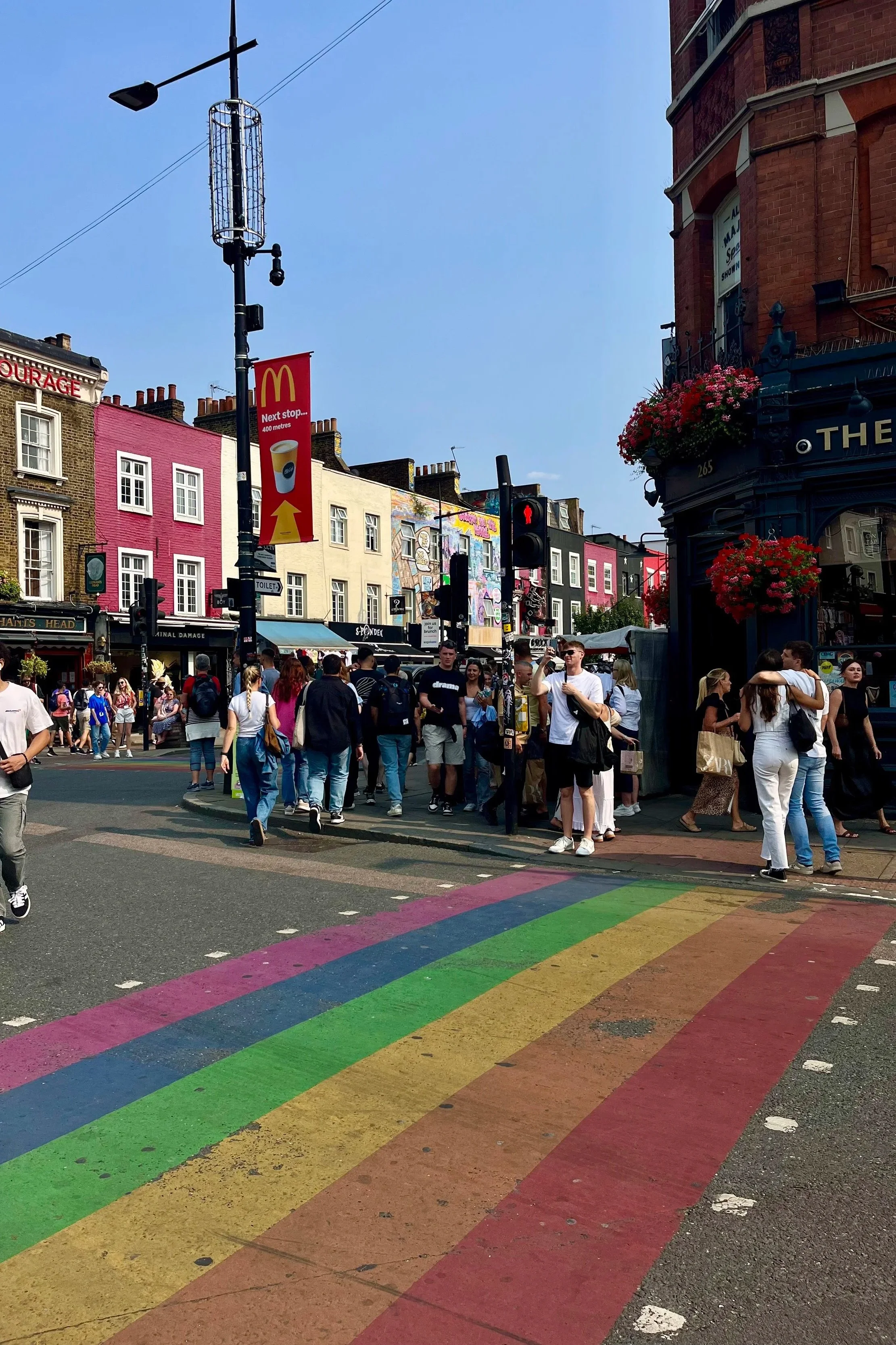 Strada pedonale con passanti e incrocio pedonale con strisce arcobaleno, edifici colorati e persone che aspettano di attraversare, con insegne e fiori decorativi. Ci troviamo a Camden Market.